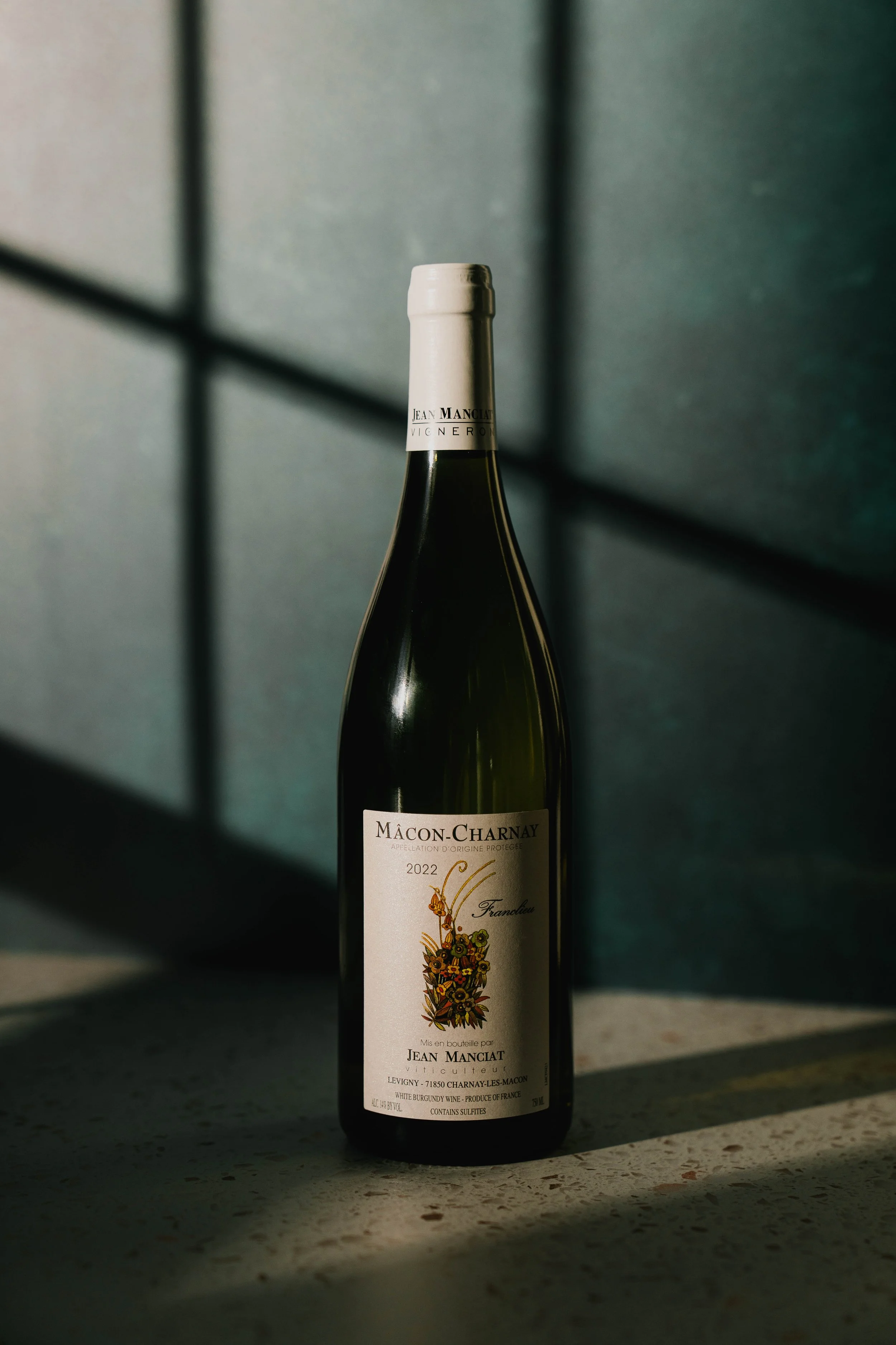 A bottle of white wine with a white label and silver cap, placed on a table with a dark, textured background.