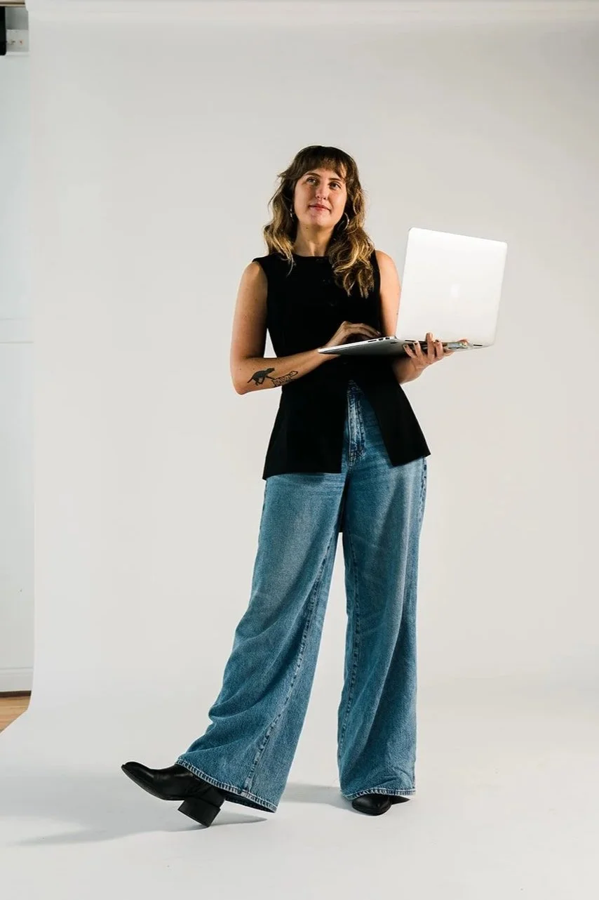 A woman with wavy hair wearing a sleeveless black top, wide-leg jeans, and black shoes, standing and holding a silver laptop against a plain background.