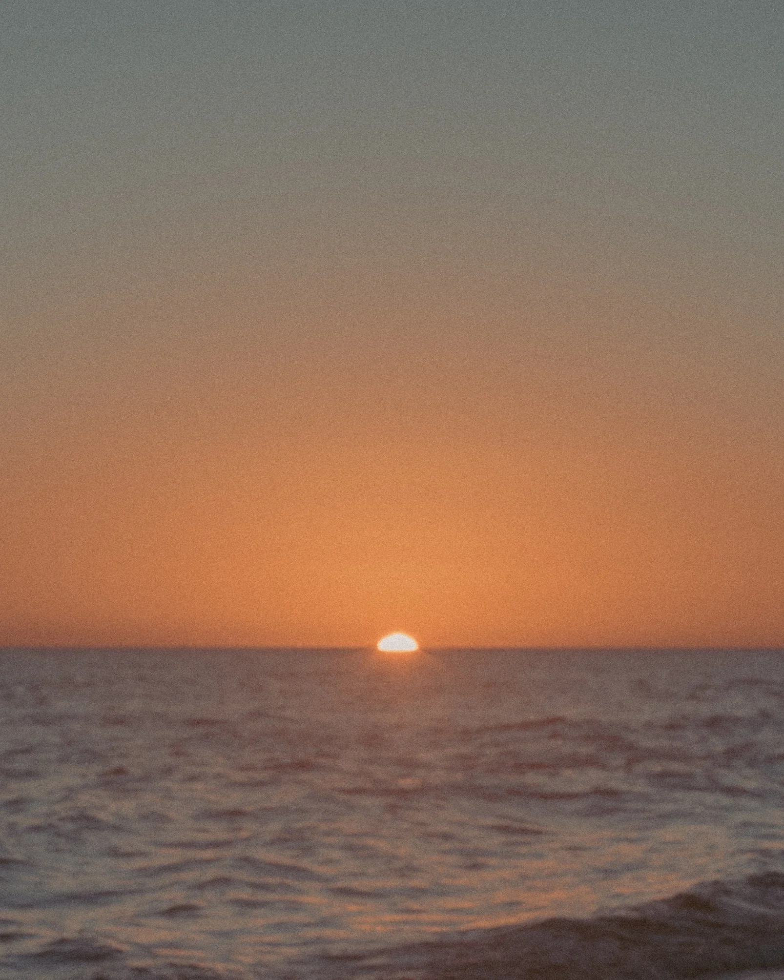 Sunset over the ocean with the sun partially below the horizon and the sky in shades of orange, fading to gray, with water in the foreground
