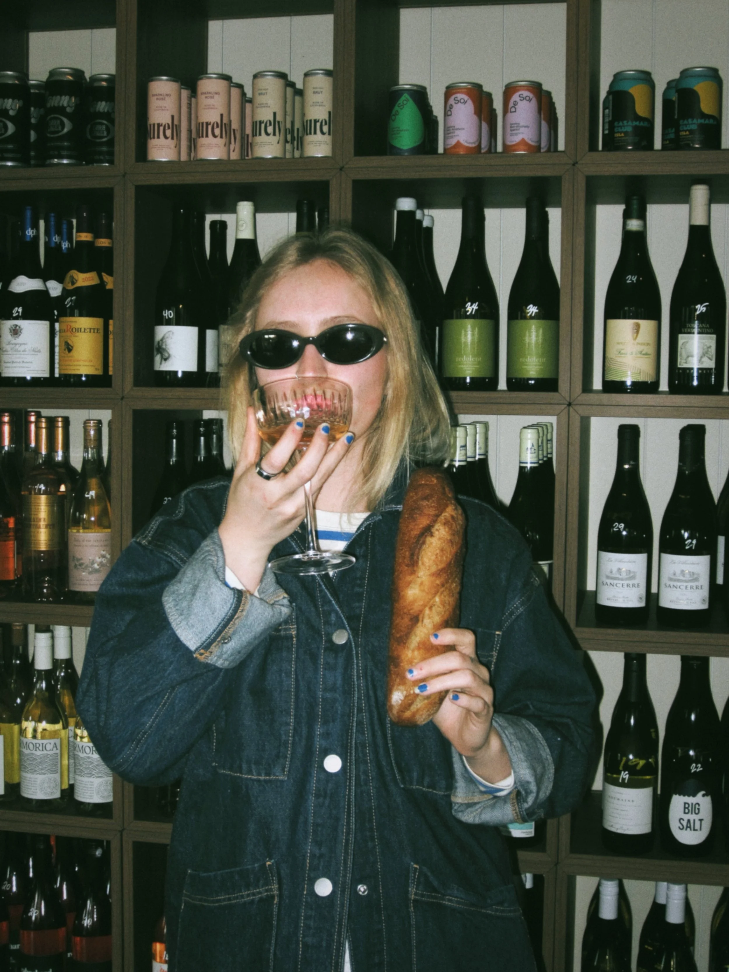 A person wearing sunglasses, holding a glass of wine in one hand and a baguette in the other, standing in front of a wine shelf.