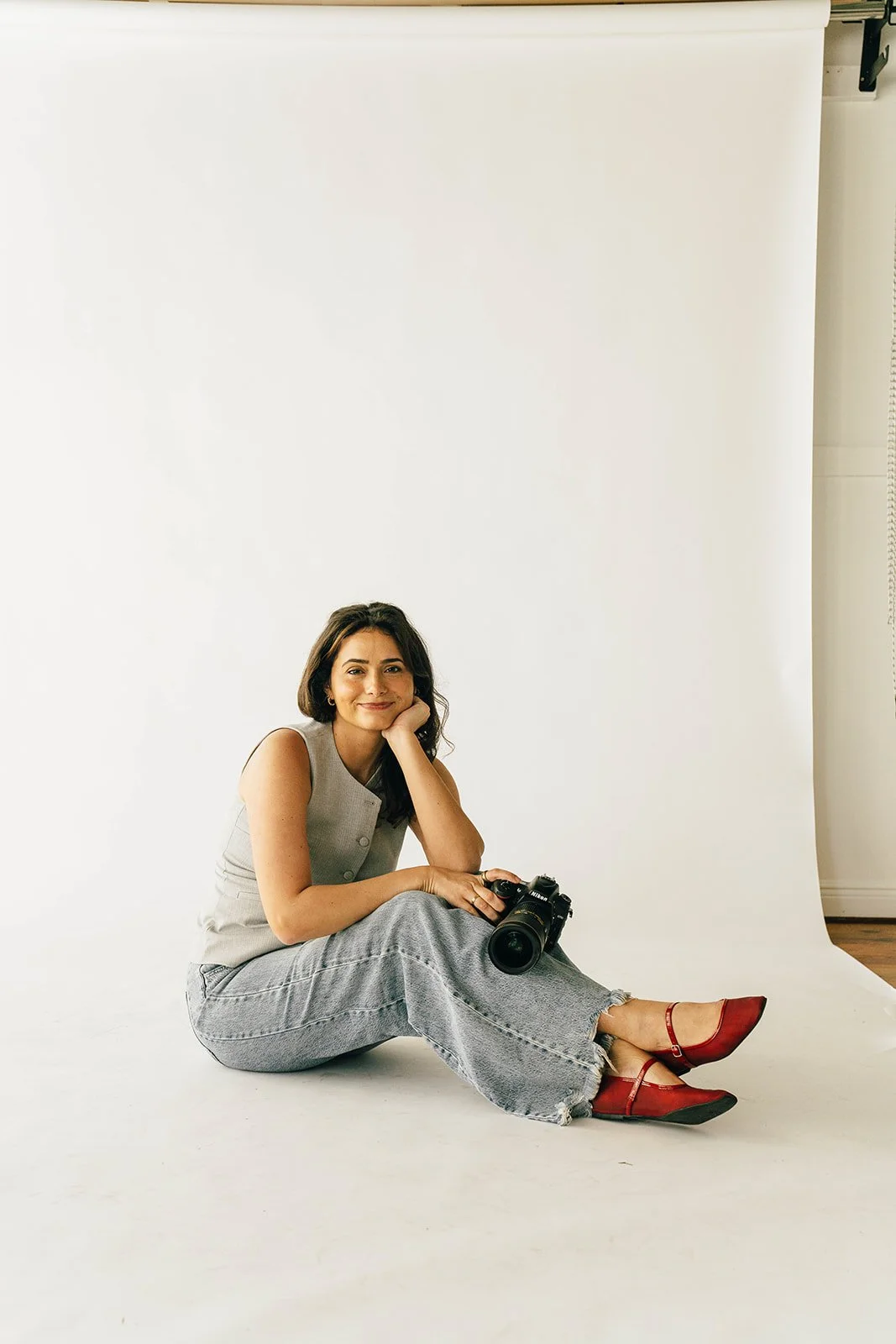 A woman sitting on the floor in front of a white backdrop, holding a camera, wearing a sleeveless top, wide-leg jeans, and red shoes.
