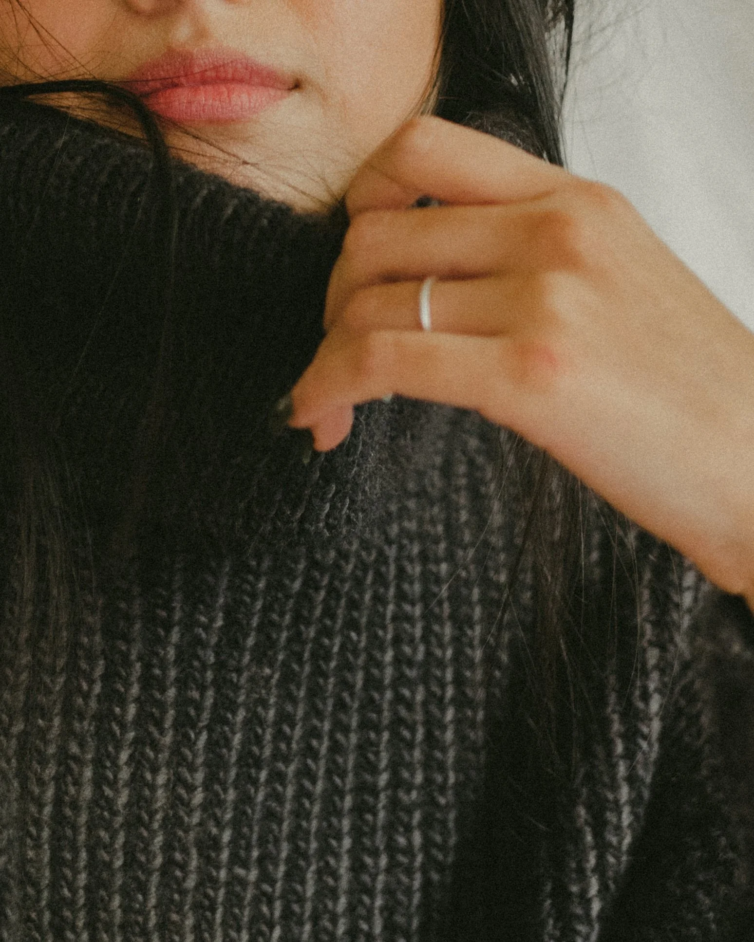 Close-up of a woman wearing a black and gray knit sweater, partially covering her face with her hand resting near her chin. She has long dark hair and a ring on her finger.