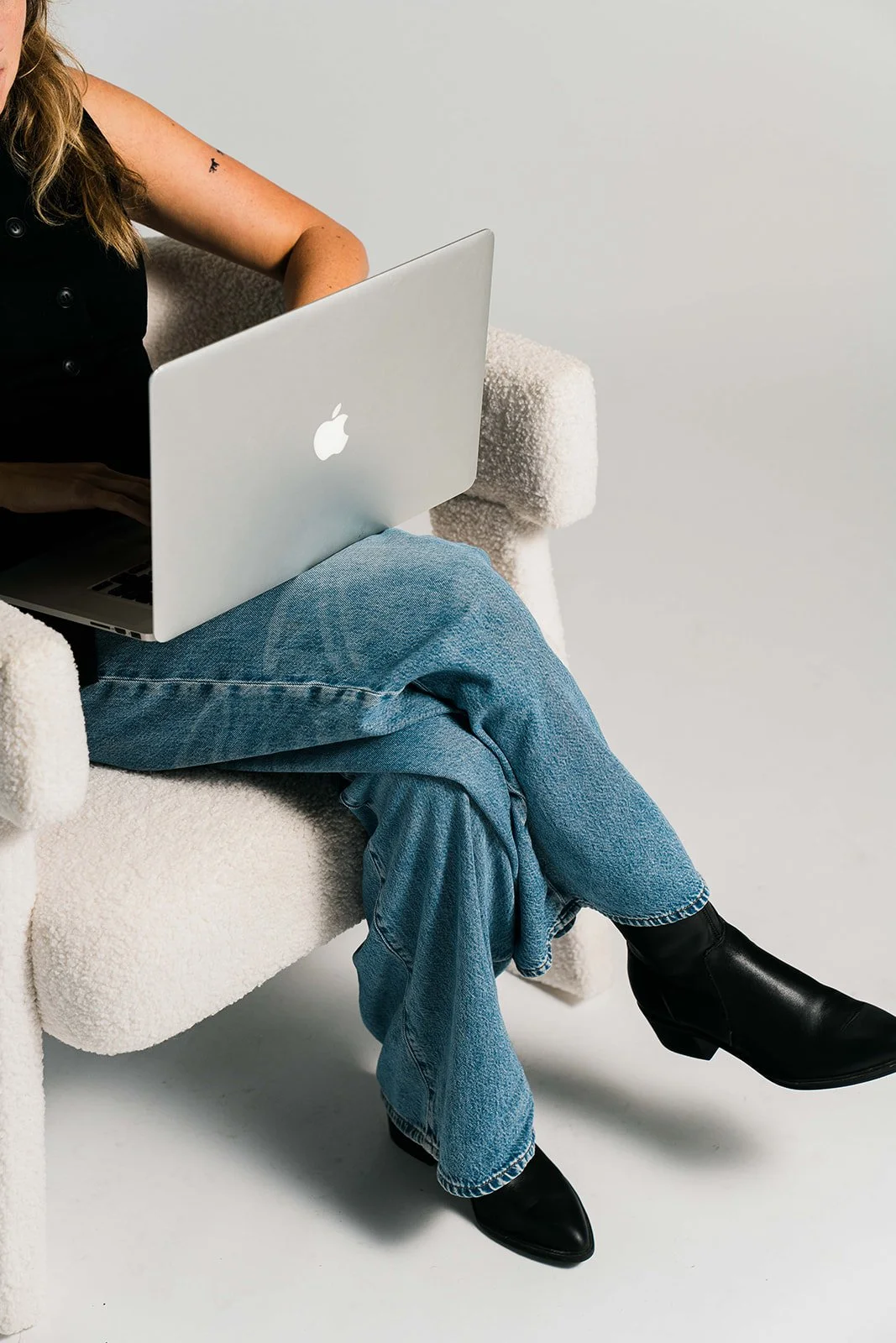 A woman sitting on a fluffy white chair using a silver MacBook, dressed in a black top, blue jeans, and black ankle boots.