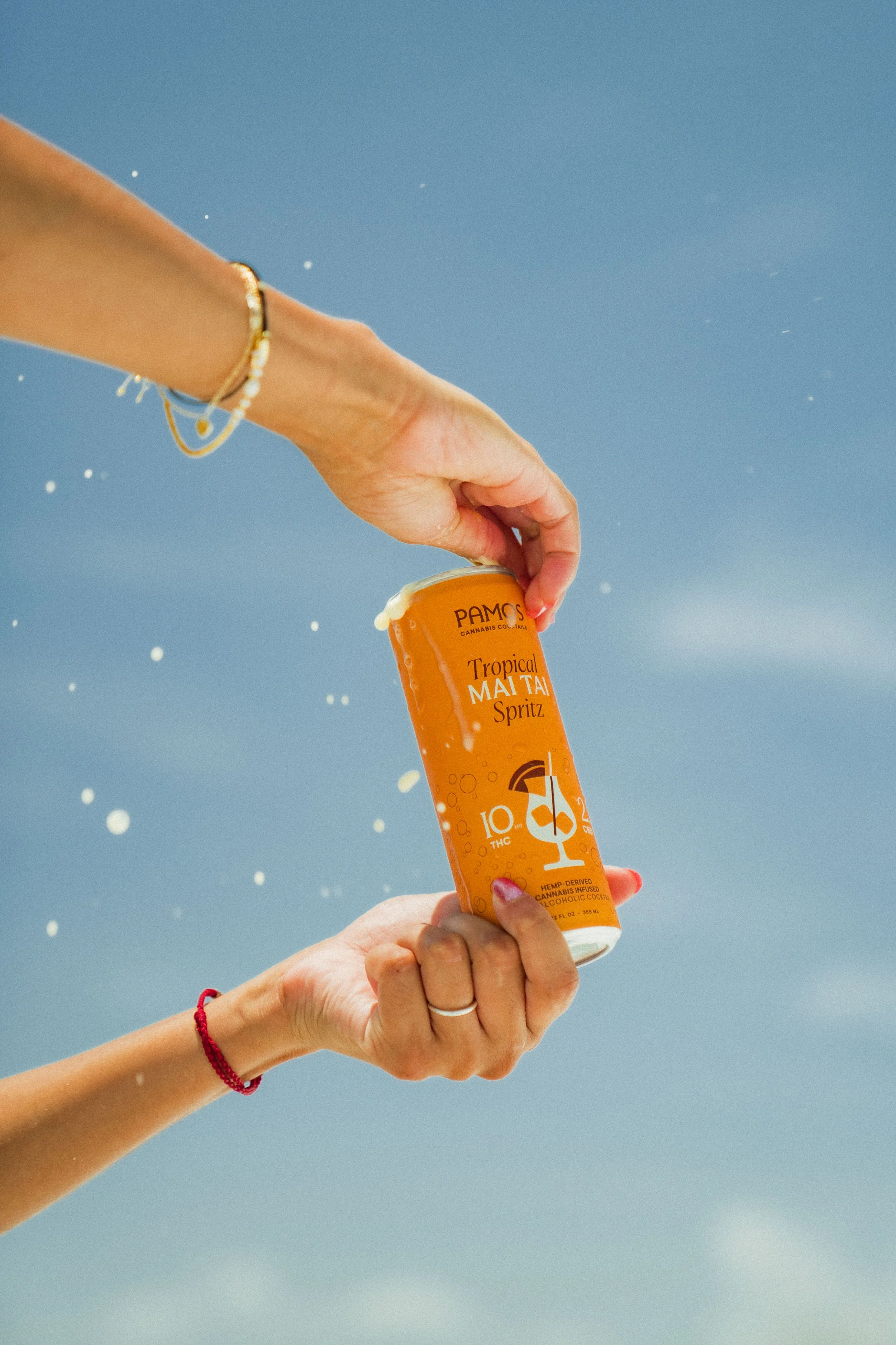 Two hands exchanging a can of Pamos Tropical Mai Tai Spritz with a blue sky background
