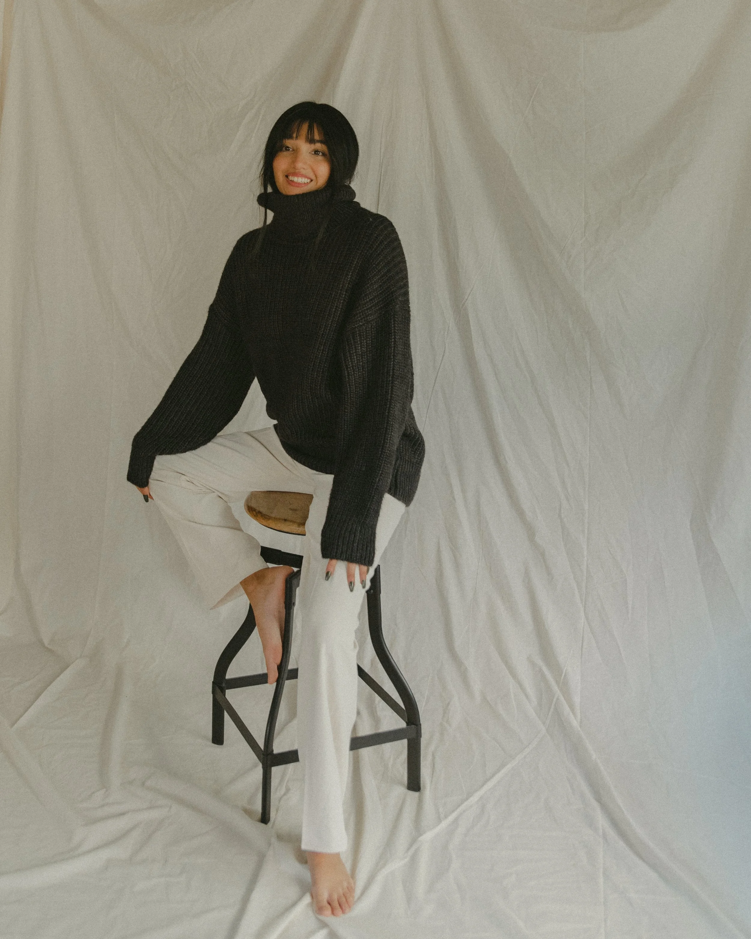 A woman with black hair wearing a black turtleneck sweater and white pants, sitting on a stool in front of a beige backdrop, smiling.