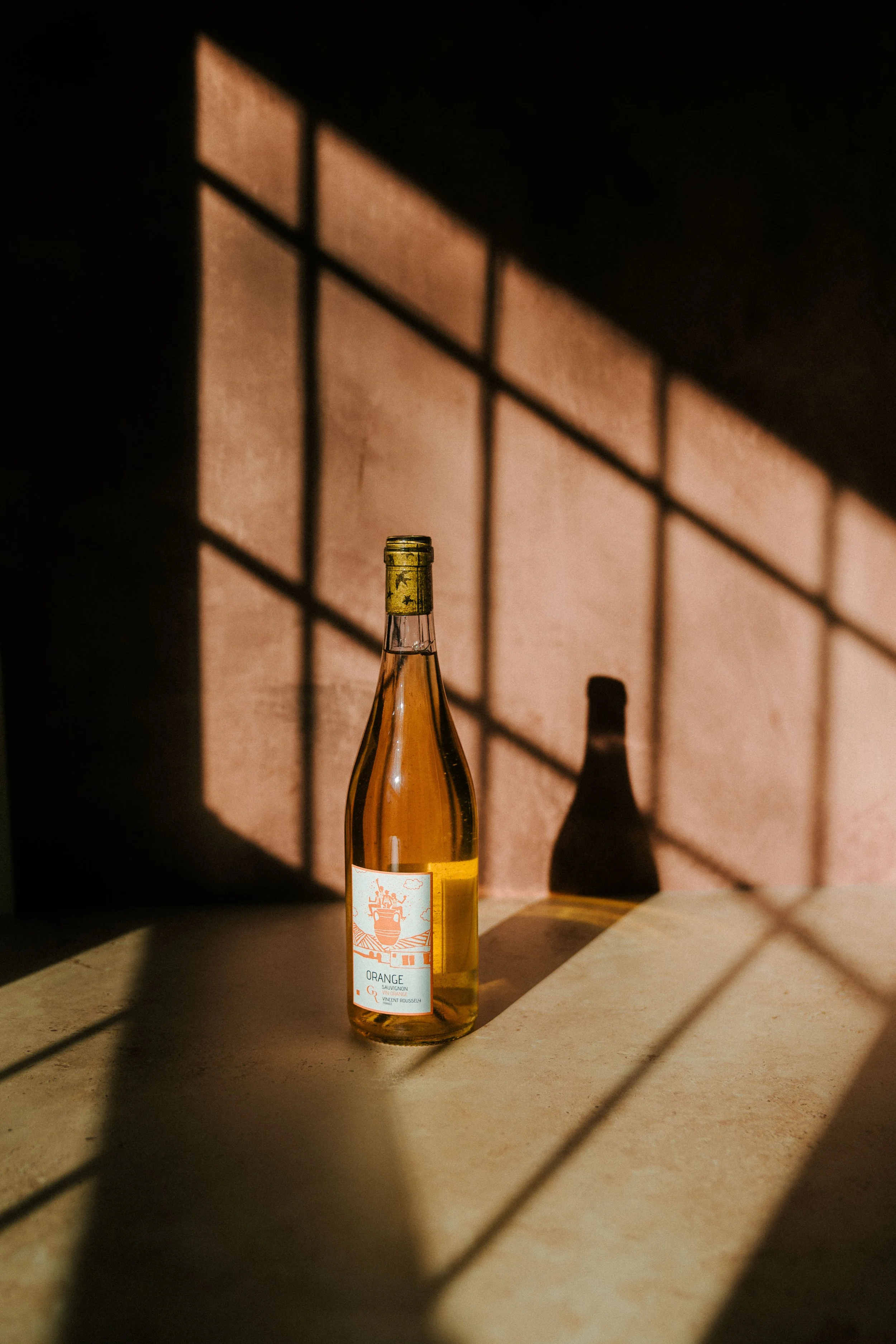 A bottle of orange wine placed on a beige surface with shadows cast by a window grid pattern on the wall in the background.