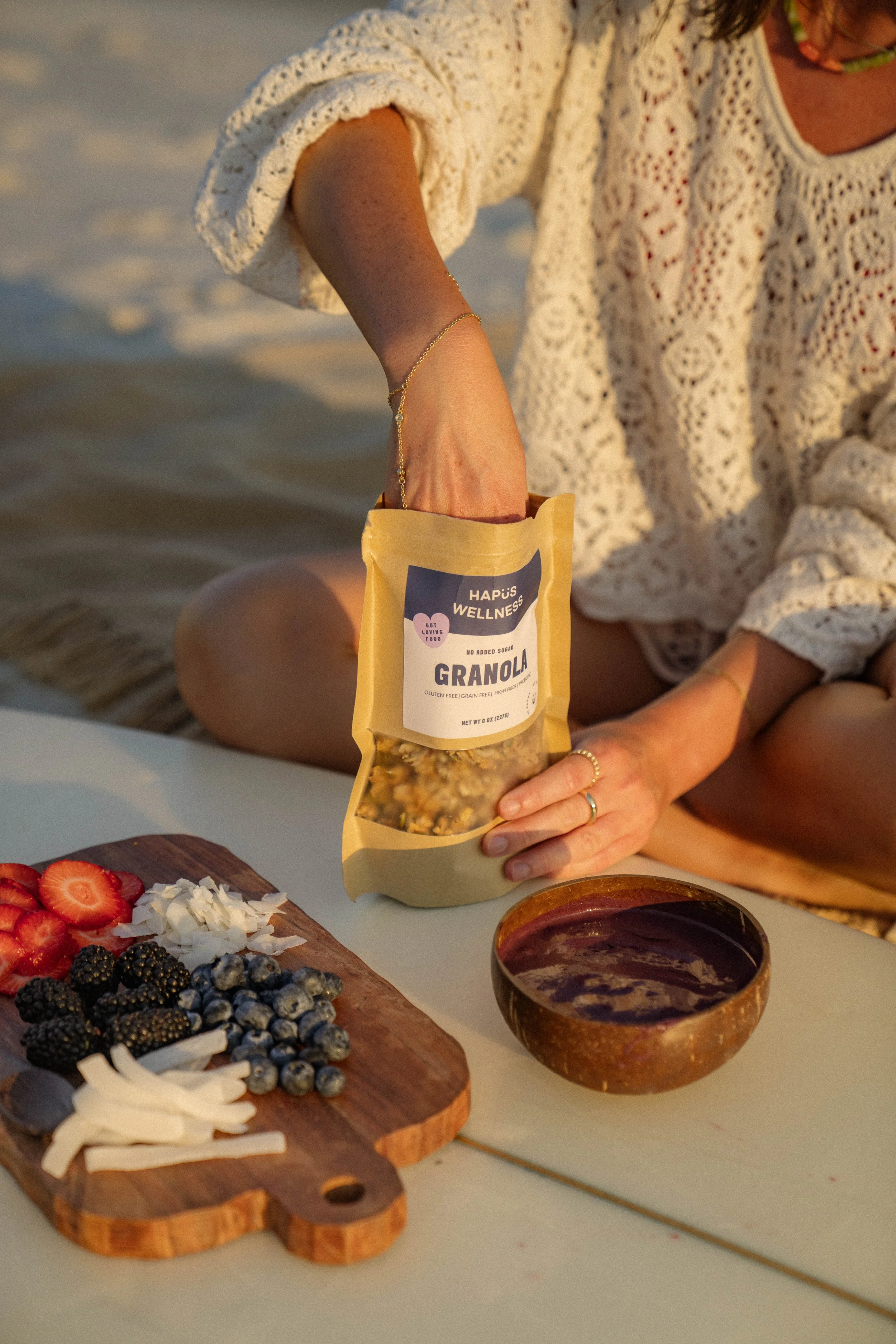 A woman sitting on the beach with a wooden board of sliced strawberries, blackberries, blueberries, and coconut flakes, and a bowl of dark sauce, while she reaches into a bag of granola.