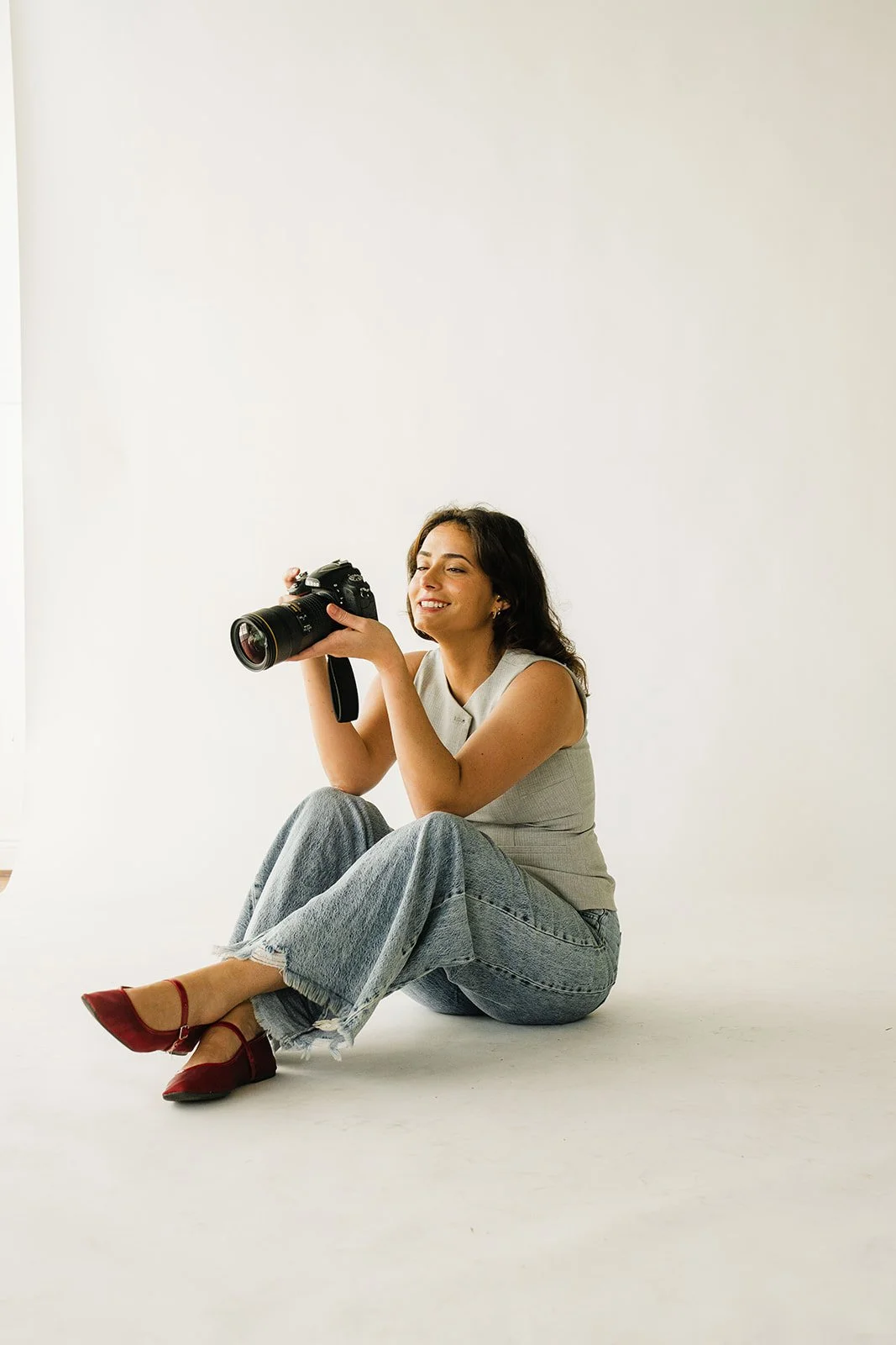 A woman sitting on the floor, smiling and holding a camera, against a plain white background.