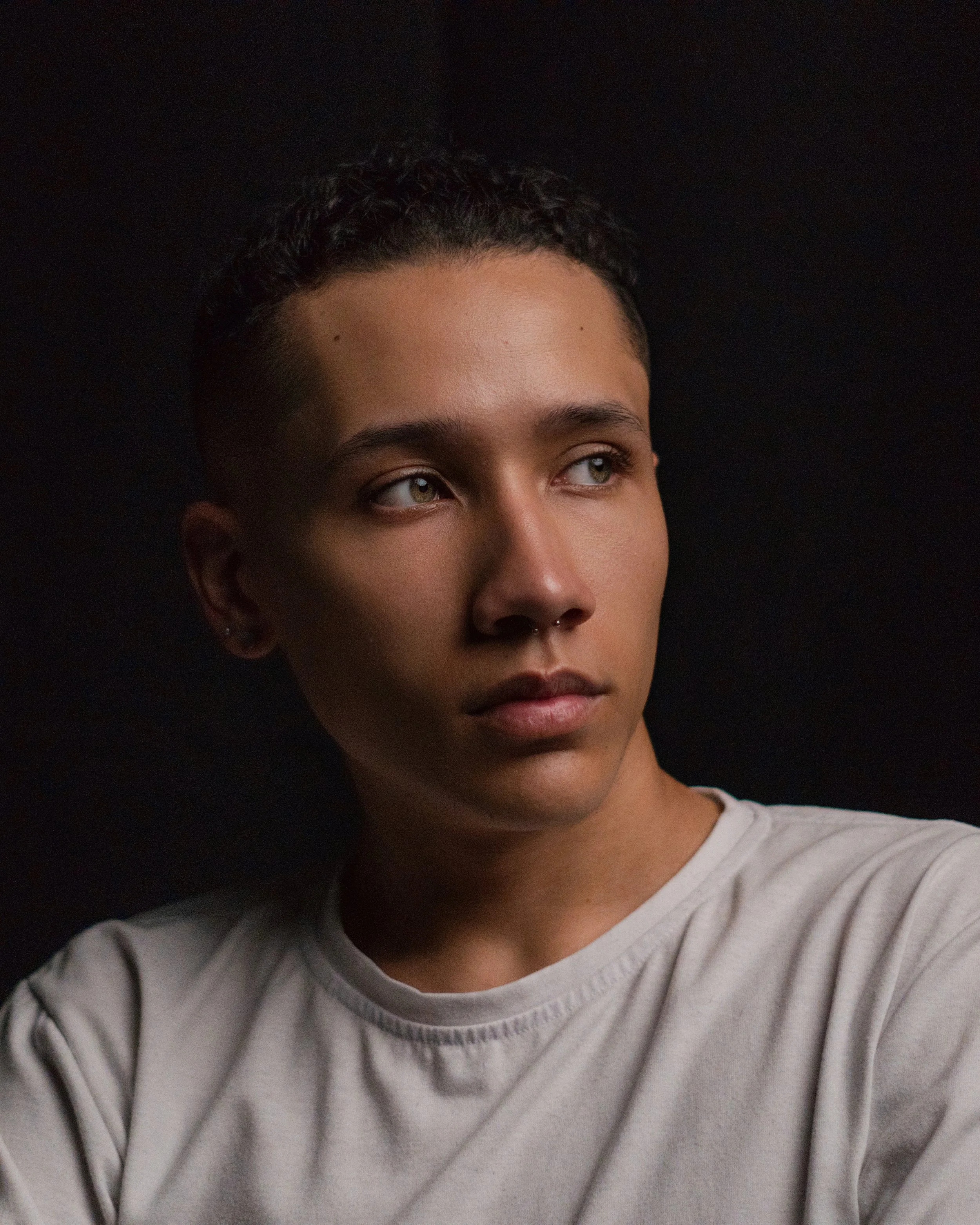 Portrait of a young person with short dark hair, light skin, green eyes, wearing a white t-shirt, against a dark background.