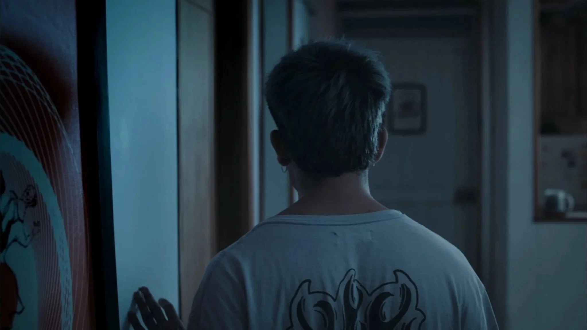 Back view of a person with short hair wearing a white t-shirt, standing in a dimly lit room, facing a wall with a framed picture.