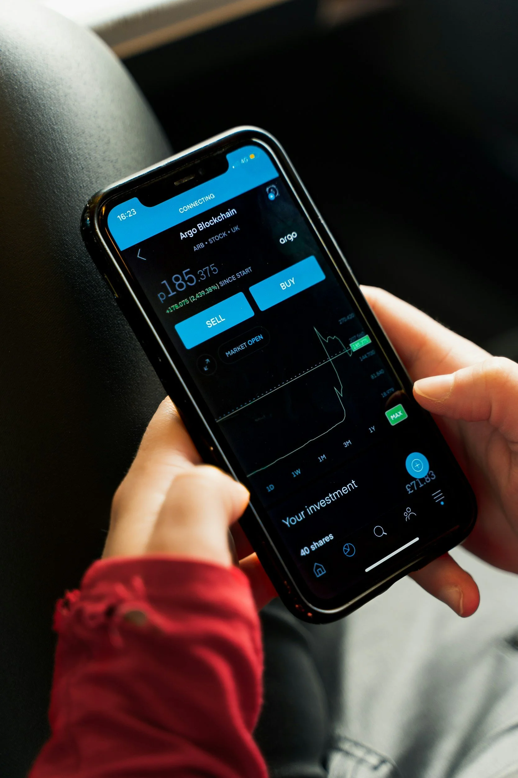 Person holding a smartphone displaying a cryptocurrency trading app for Argo Blockchain, showing the current price, buy and sell options, and a price chart.
