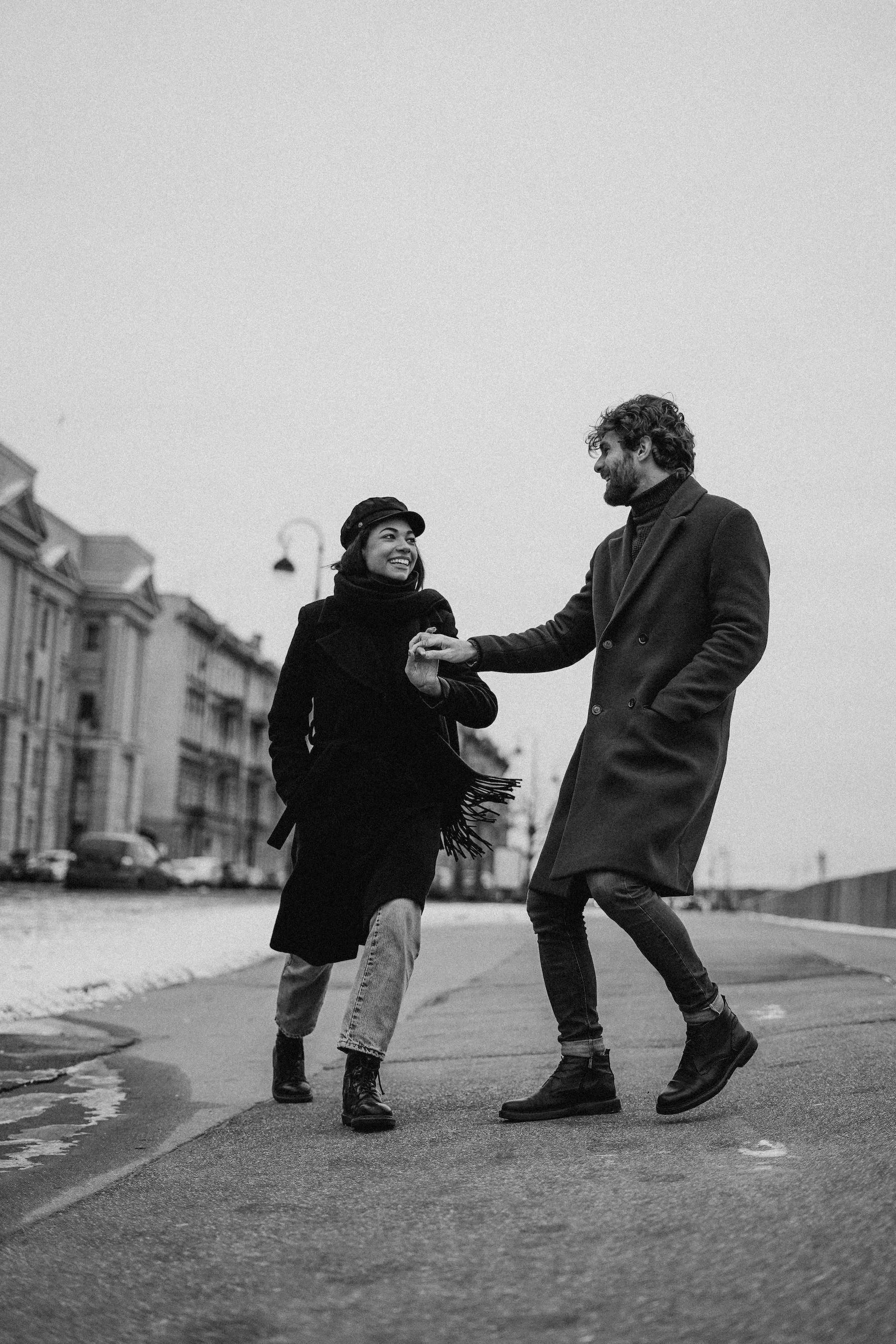 A black-and-white photo of a couple holding hands and dancing on a street, smiling at each other, with overcast sky and residential buildings in the background.