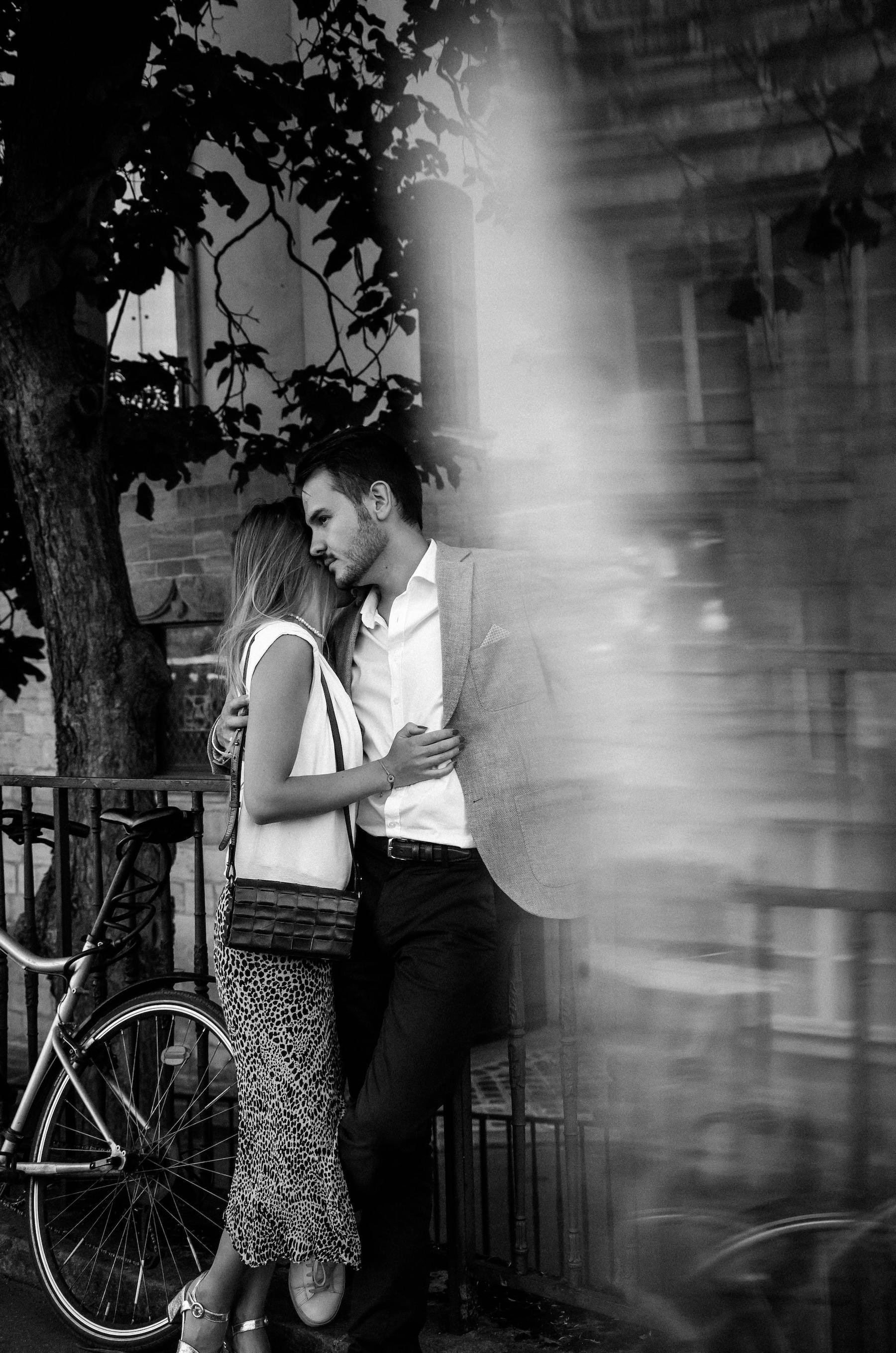 A black-and-white photo of a couple embracing on a city street. The woman has long hair and is wearing a sleeveless top, patterned pants, and high heels, with a purse over her shoulder. The man has short hair and is dressed in a blazer, white shirt, and dark pants. They are standing next to a bicycle with a tree and building in the background.