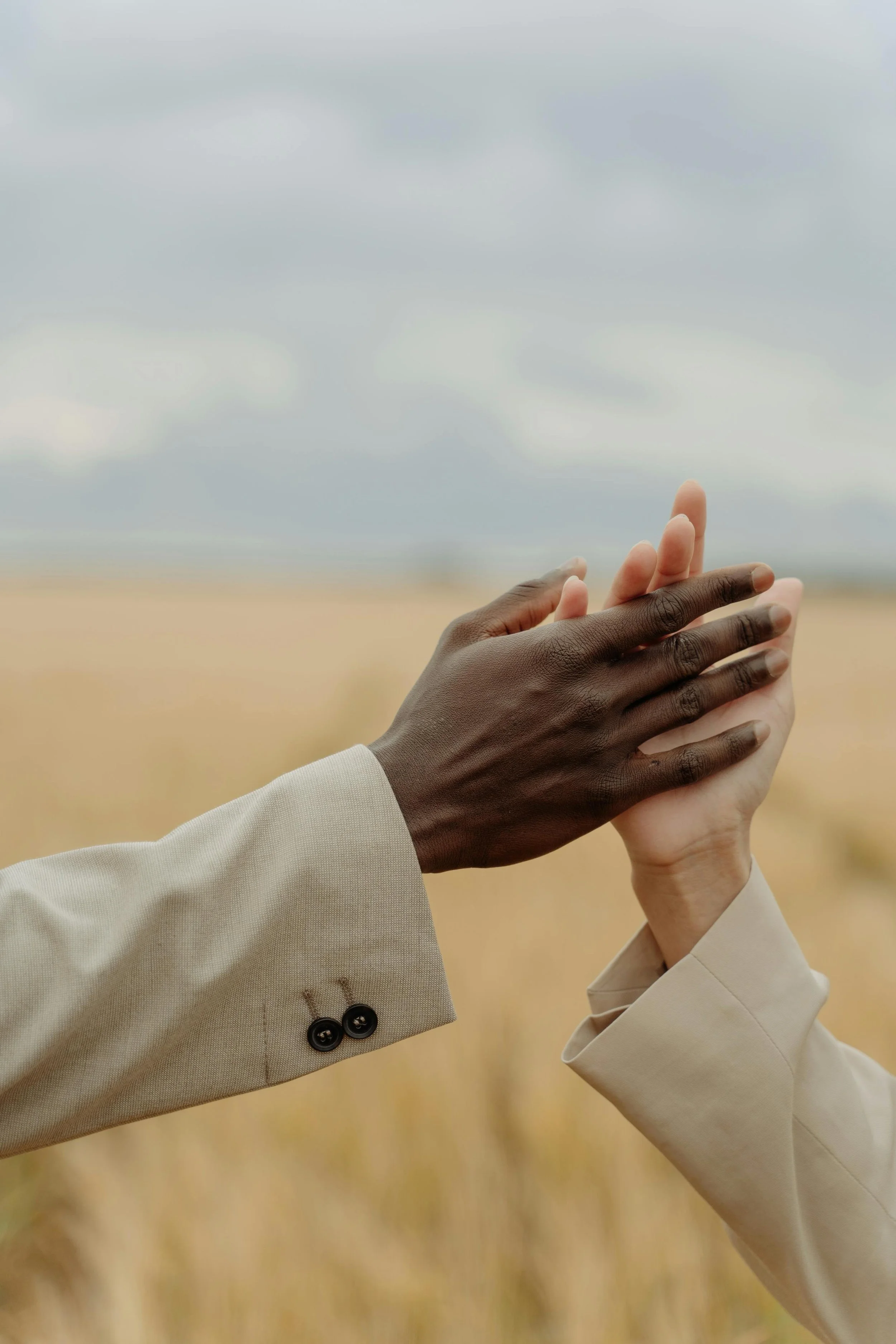 Two hands, one dark-skinned and one light-skinned, clasped together against a blurred outdoor background with cloudy sky, both wearing beige jackets.