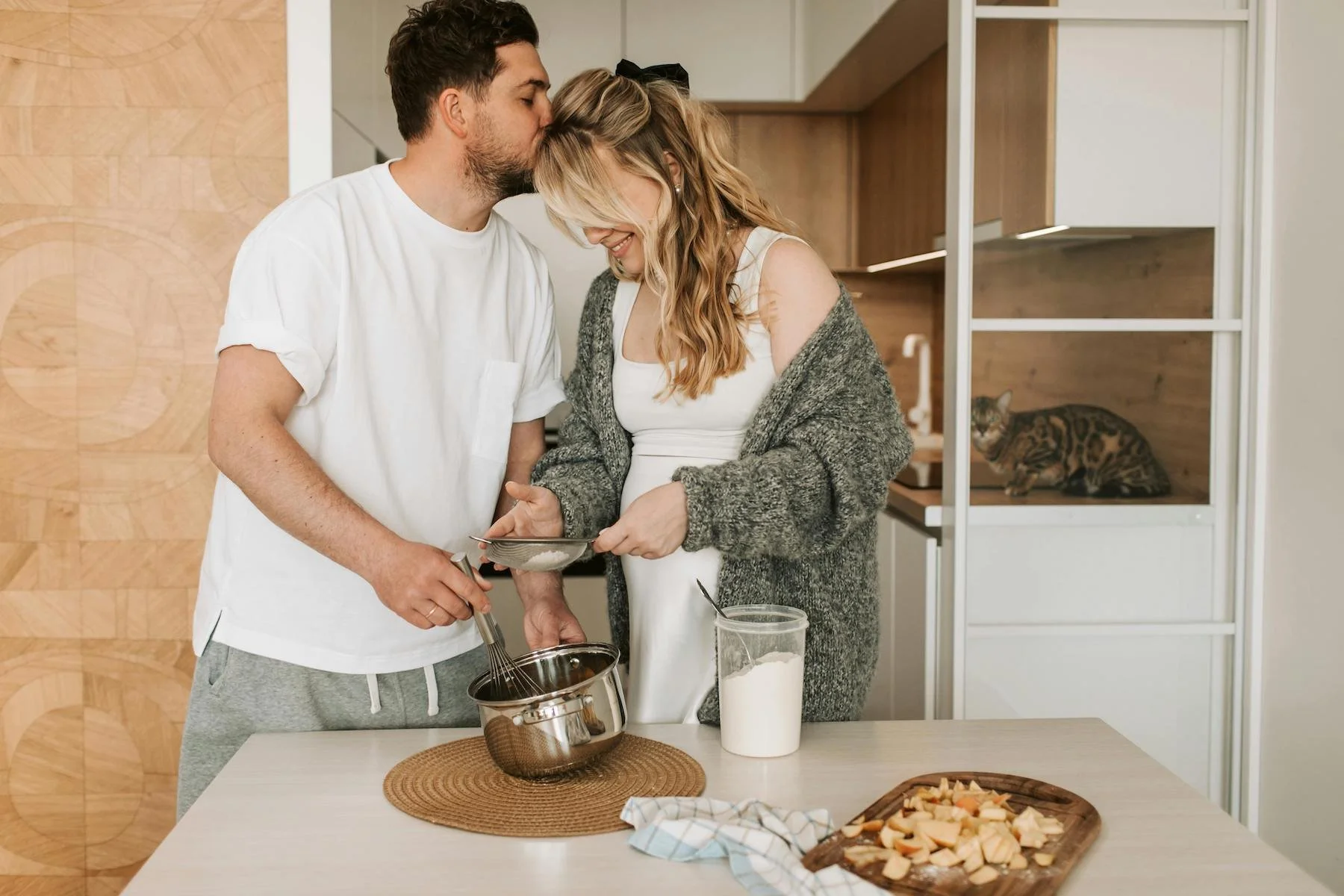 A young couple in a kitchen preparing to bake together, with a cat resting inside a white wooden cabinet in the background.