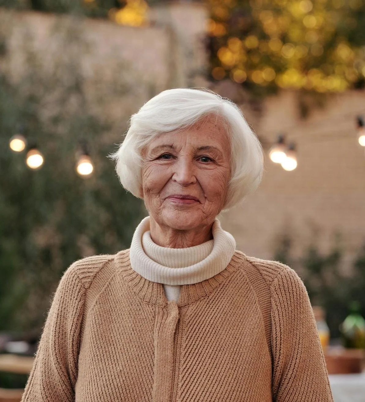 Older woman with white hair smiling outdoors during evening or late afternoon, wearing a beige knitted sweater with a turtleneck.