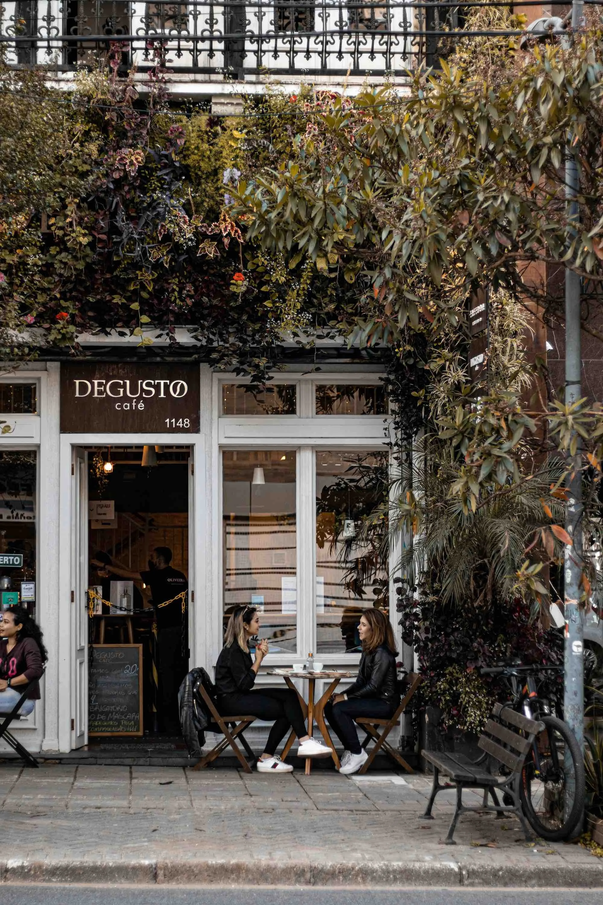 Two women are sitting at a small table outside a coffee shop called Degusto Café, engaging in conversation. There is greenery and plants surrounding the café, and a bicycle is parked nearby.
