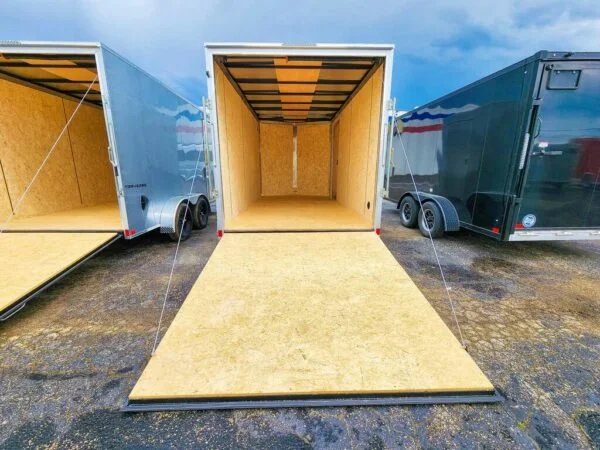 Three enclosed cargo trailers with open doors, showing empty interior and ramp doors for loading, parked on a gravel lot.
