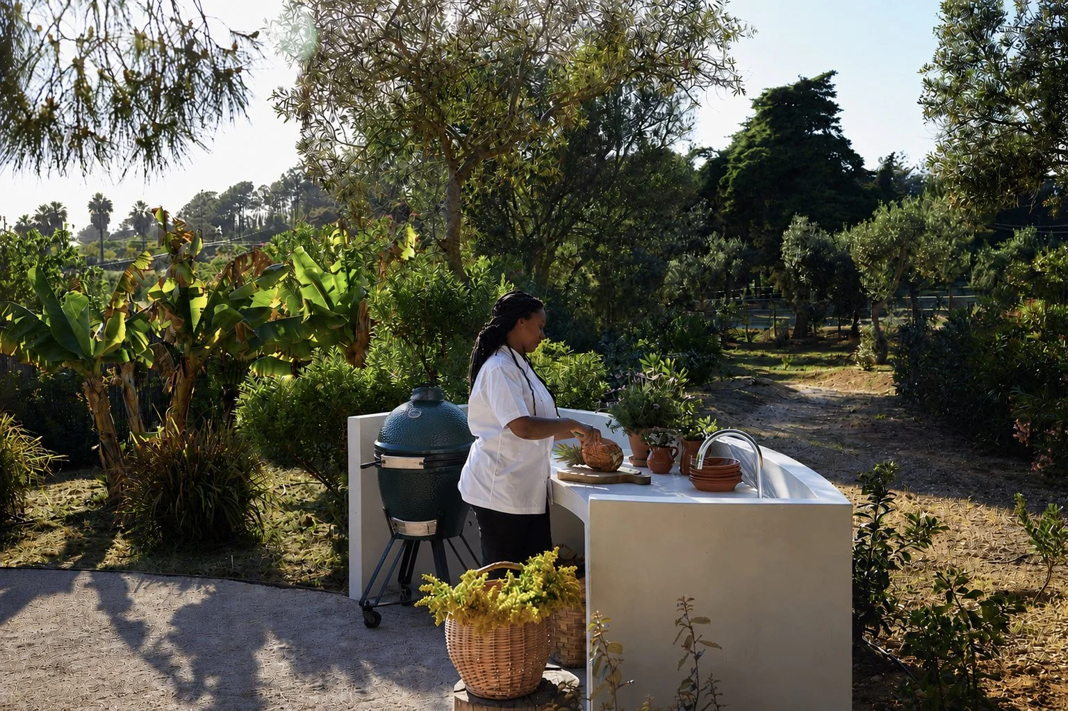A woman in a white shirt standing at an outdoor white kitchen counter in a garden, preparing food. The scene is sunny with lush green trees and plants surrounding her.