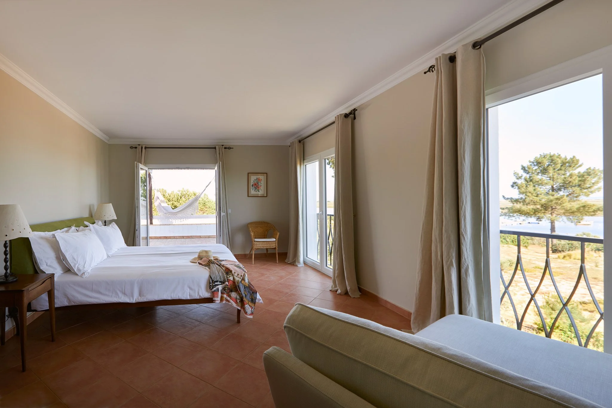 Spacious hotel room with a large bed, a balcony door with a hammock outside, sliding glass doors to a view of trees and water, and light-colored curtains.
