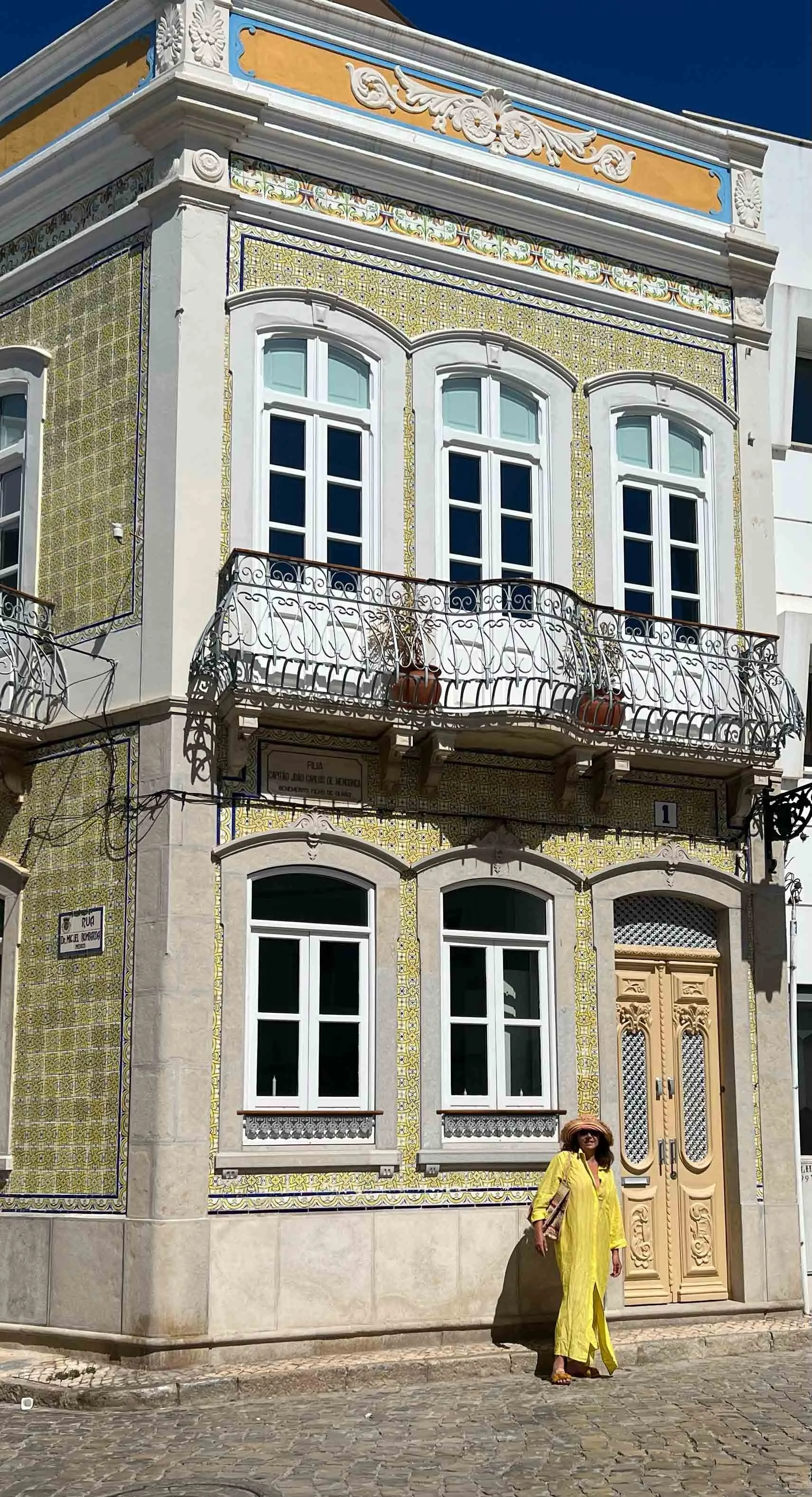 A woman dressed in yellow standing in front of a decorative, historic building with ornate tiles, elaborately carved double doors, and black wrought iron balcony railings.