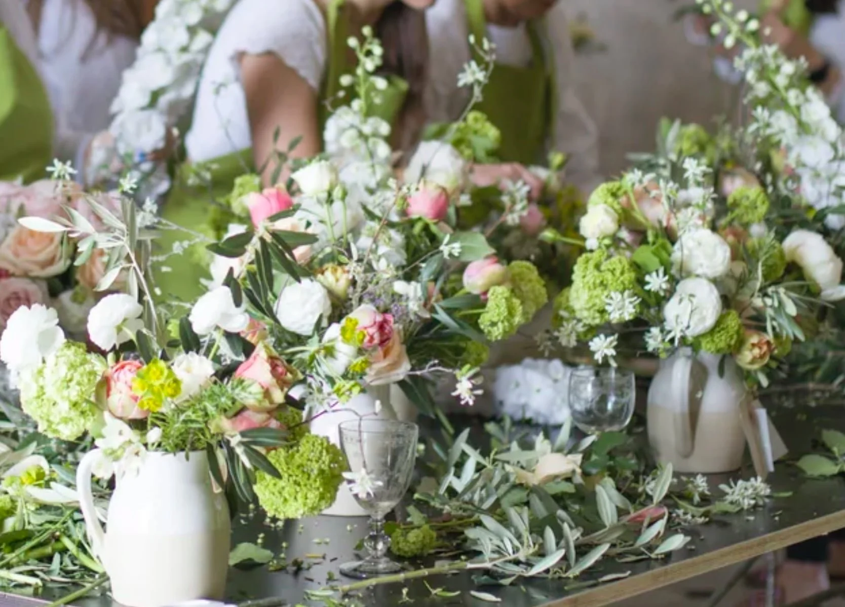Various flower arrangements in white vases on a table at a floral workshop, with some flowers and leaves scattered on the surface.