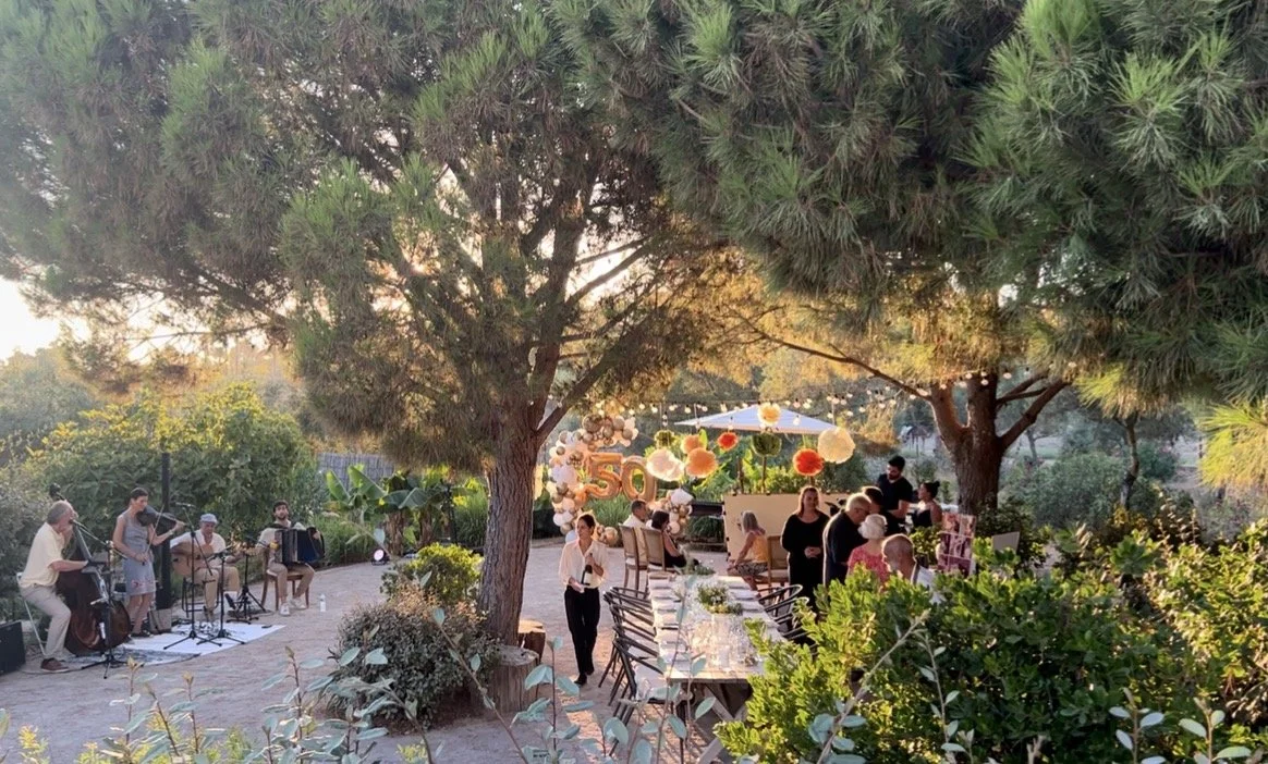 Outdoor garden party with music band, decorated with balloons and flowers, surrounded by trees and greenery, during sunset.