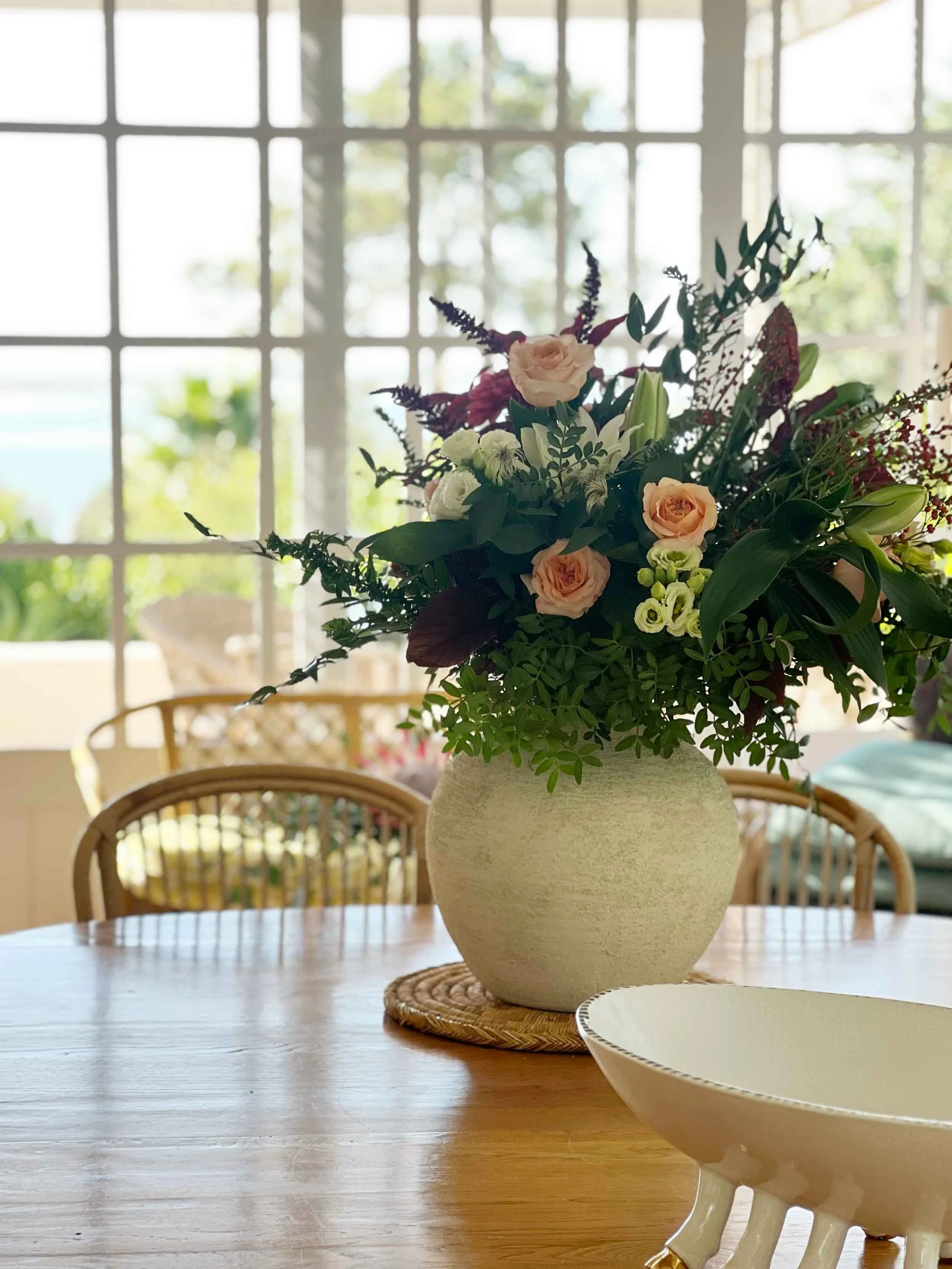 A large bouquet of flowers in a wide, off-white ceramic vase sitting on a circular mat, on a wooden table in a sunlit room with large window panes in the background.