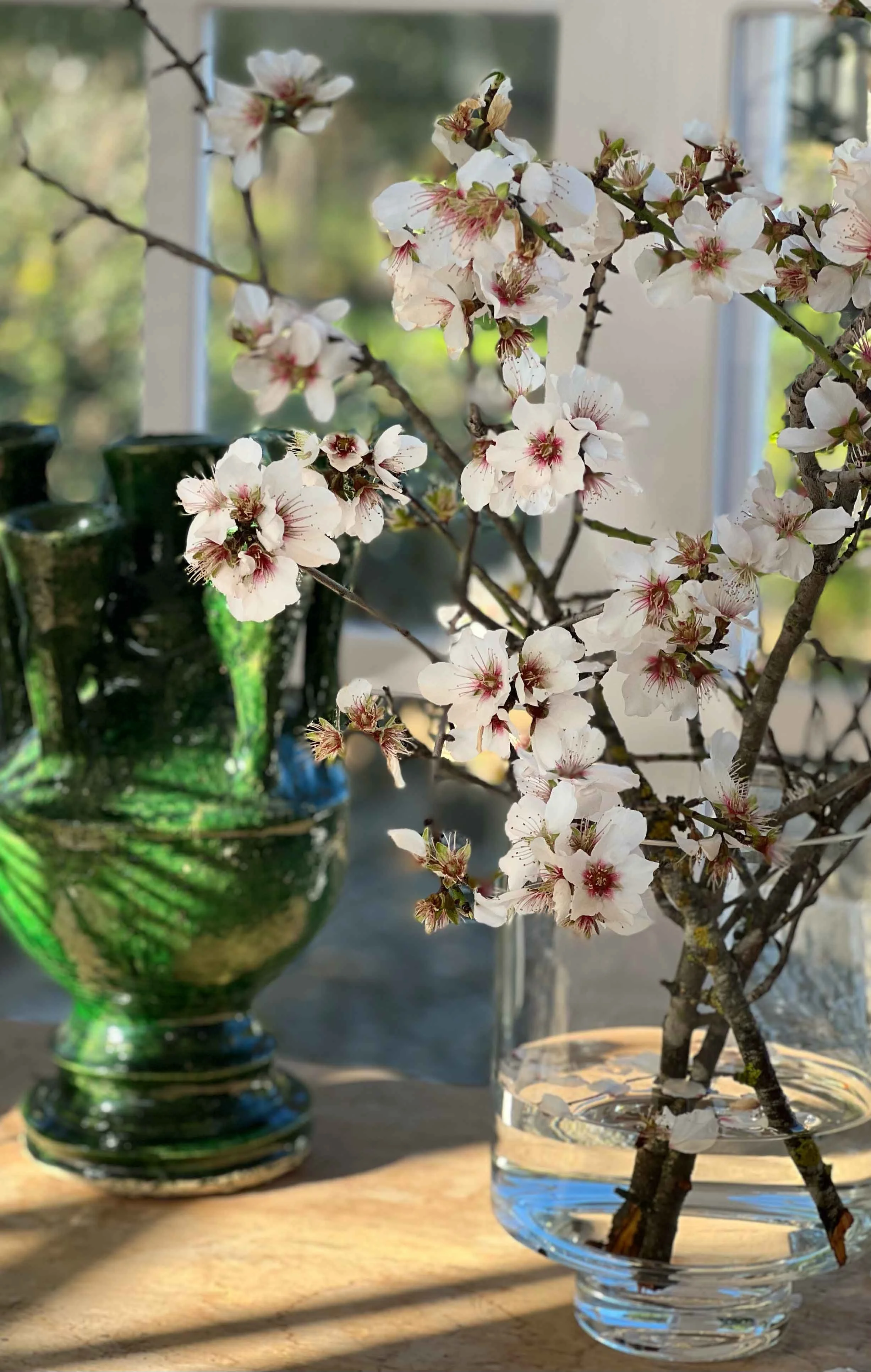 A glass vase with branches of pink and white cherry blossoms on a wooden surface, with a green glass vase and window in the background.