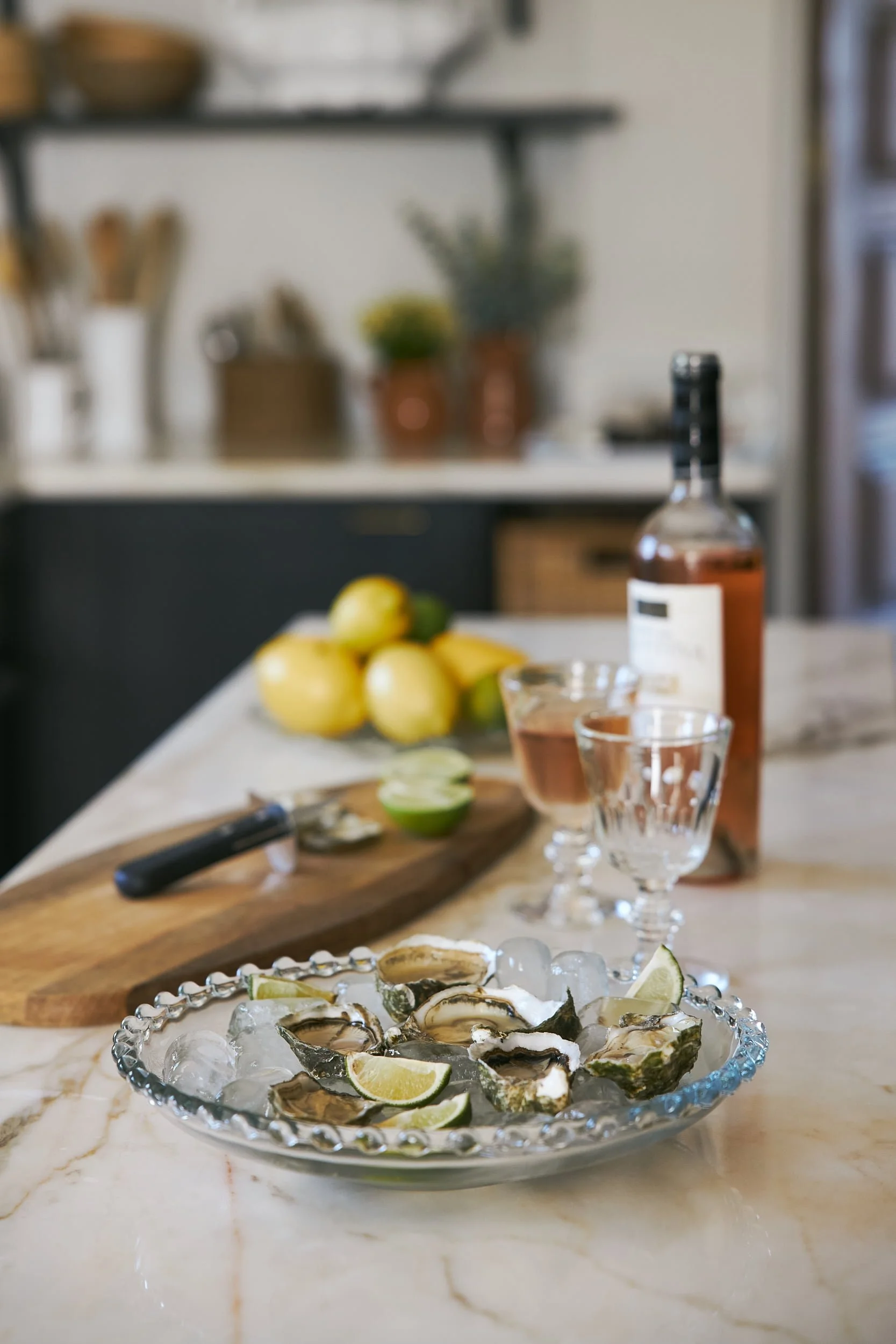 A marble kitchen countertop with a glass dish containing oysters on ice with lemon wedges, a cutting board with limes, a knife, a bottle of rosé wine, and two wine glasses, one filled with rosé wine.