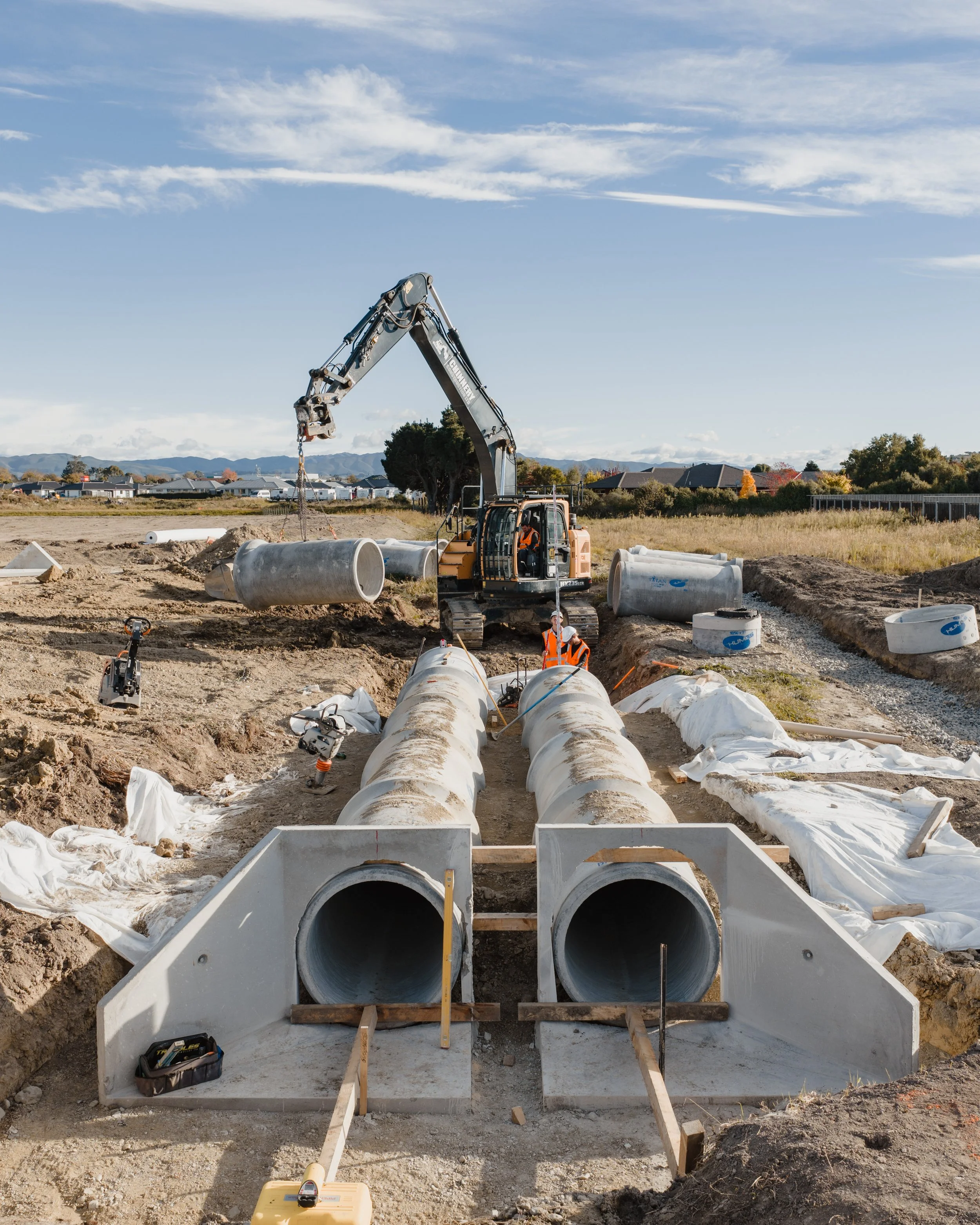 Construction site with large concrete pipes and a crane lifting pipe sections, workers in safety vests, clear sky, residential buildings in the background.