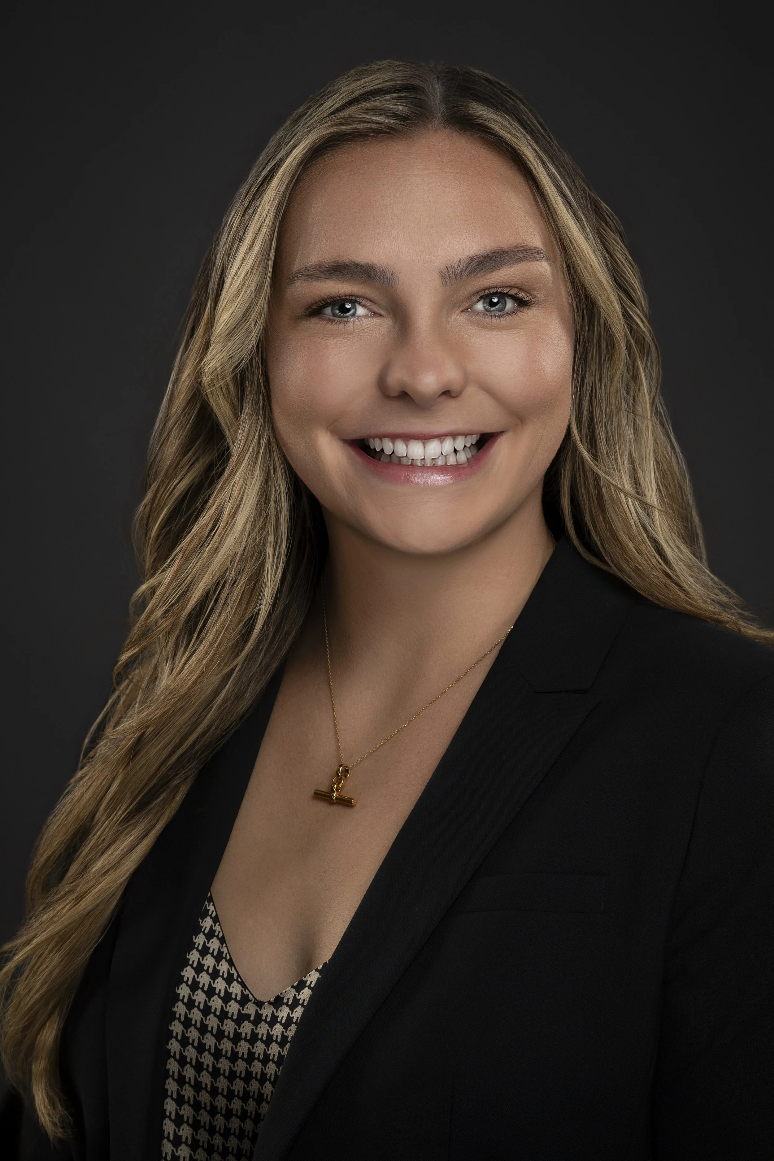 A woman with long blonde hair smiling, wearing a black blazer, a patterned blouse, and a gold necklace with a cross pendant against a dark background.