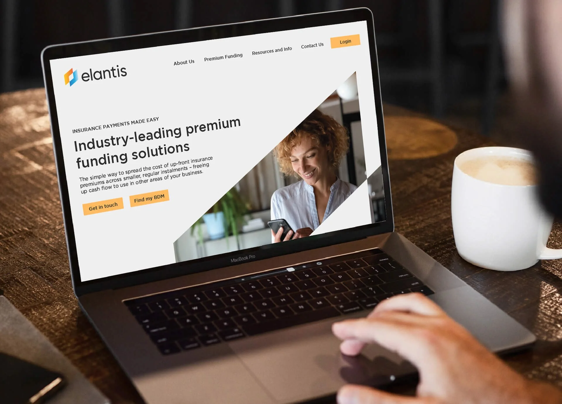 A person using a MacBook Pro laptop at a wooden table, viewing a website for Elantis that offers insurance payments and funding solutions, with a white coffee cup nearby.