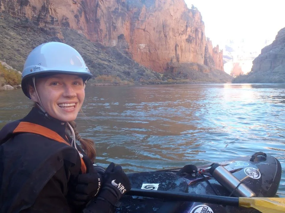 A woman in a helmet smiling while paddling a kayak on a river with canyon walls in the background.