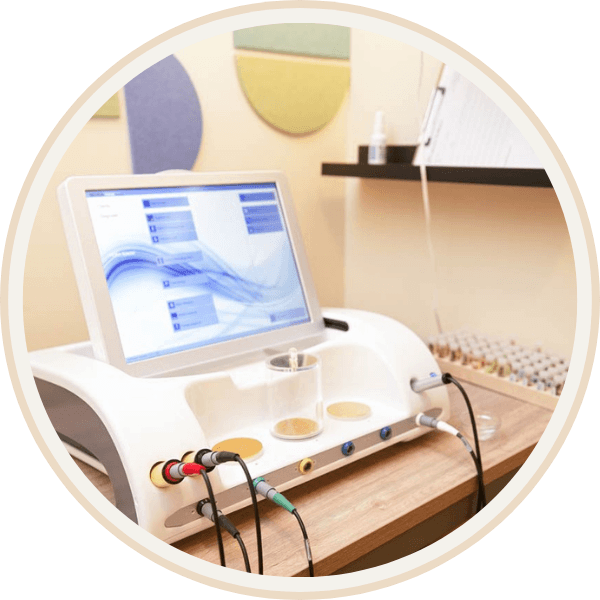 An EAV (Electro-acupuncture According to Voll) medical laboratory instrument at Current Wellness is connected to various wires on a wooden table with test tubes and colorful wall decor in the background.