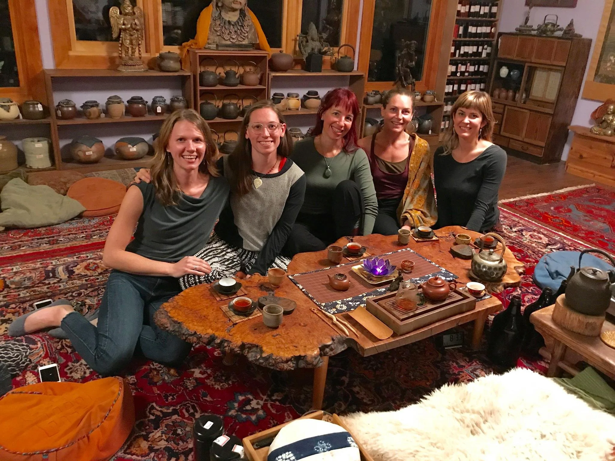 Five women sitting on a carpeted floor in front of a wooden table with tea cups, in a room decorated with pottery, statues, and wooden furniture.