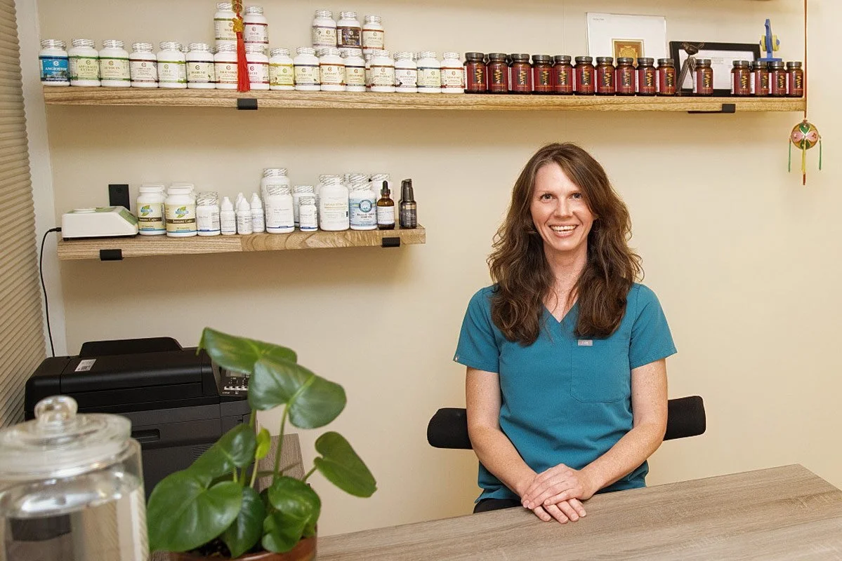 Jennifer Peterson of Current Wellness in Portland with long brown wavy hair sits, wearing blue scrubs, smiling softly, behind a desk with shelves of bottles behind her.