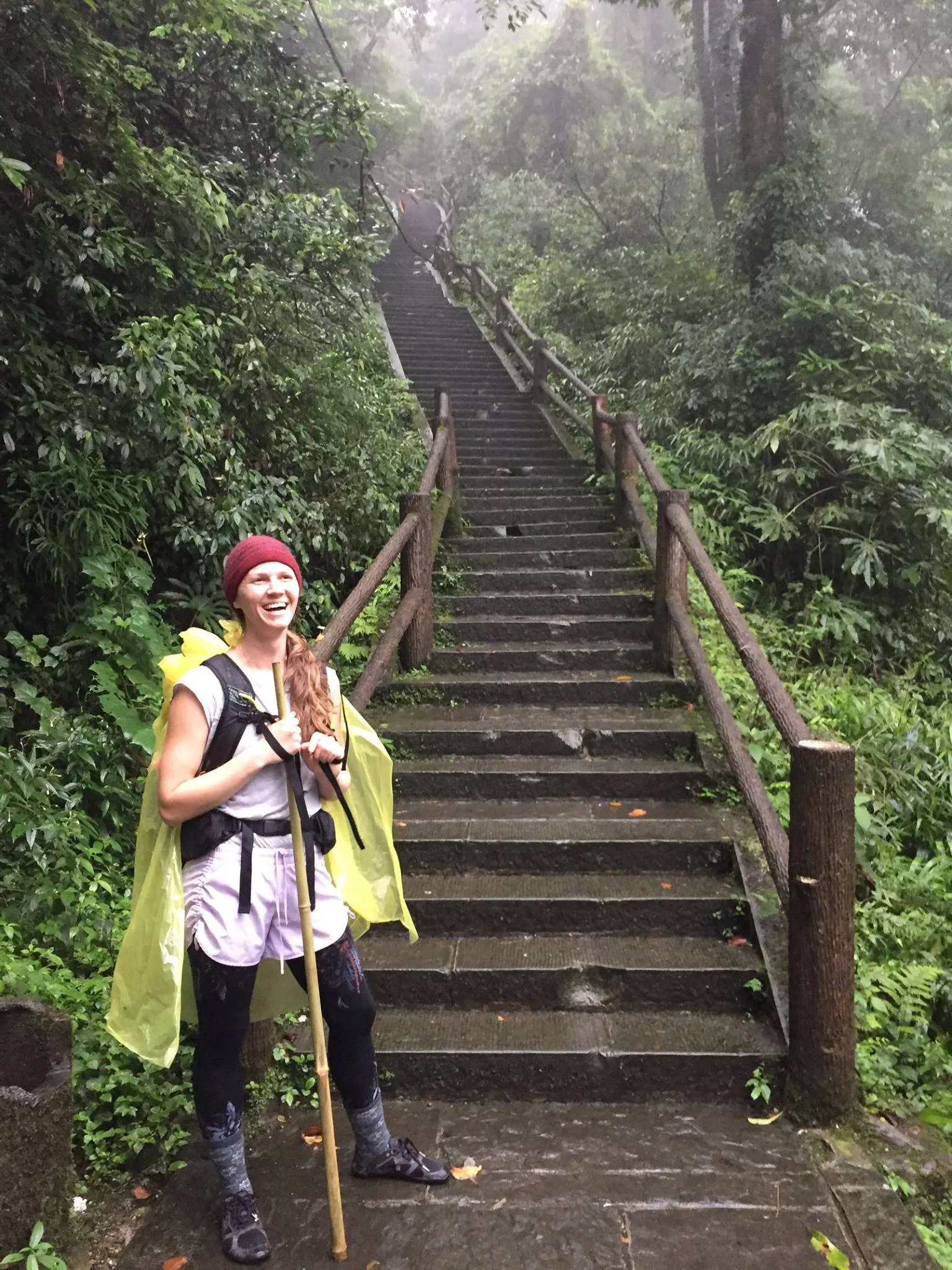 A woman with a red beanie smiling holds a hiking pole and stands at the bottom of a staircase in a lush, foggy forest. She is dressed in outdoor hiking gear, including a yellow raincoat draped over her shoulders, and has a backpack.