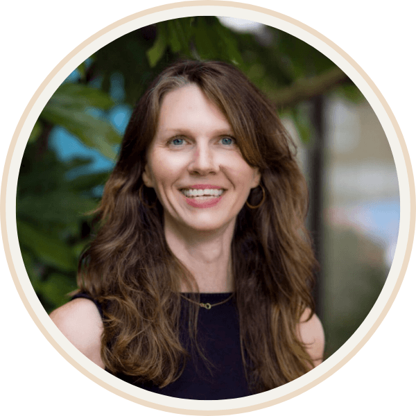 Jennifer Peterson of Current Wellness in Portland sits with long brown hair smiling outdoors in front of green foliage.