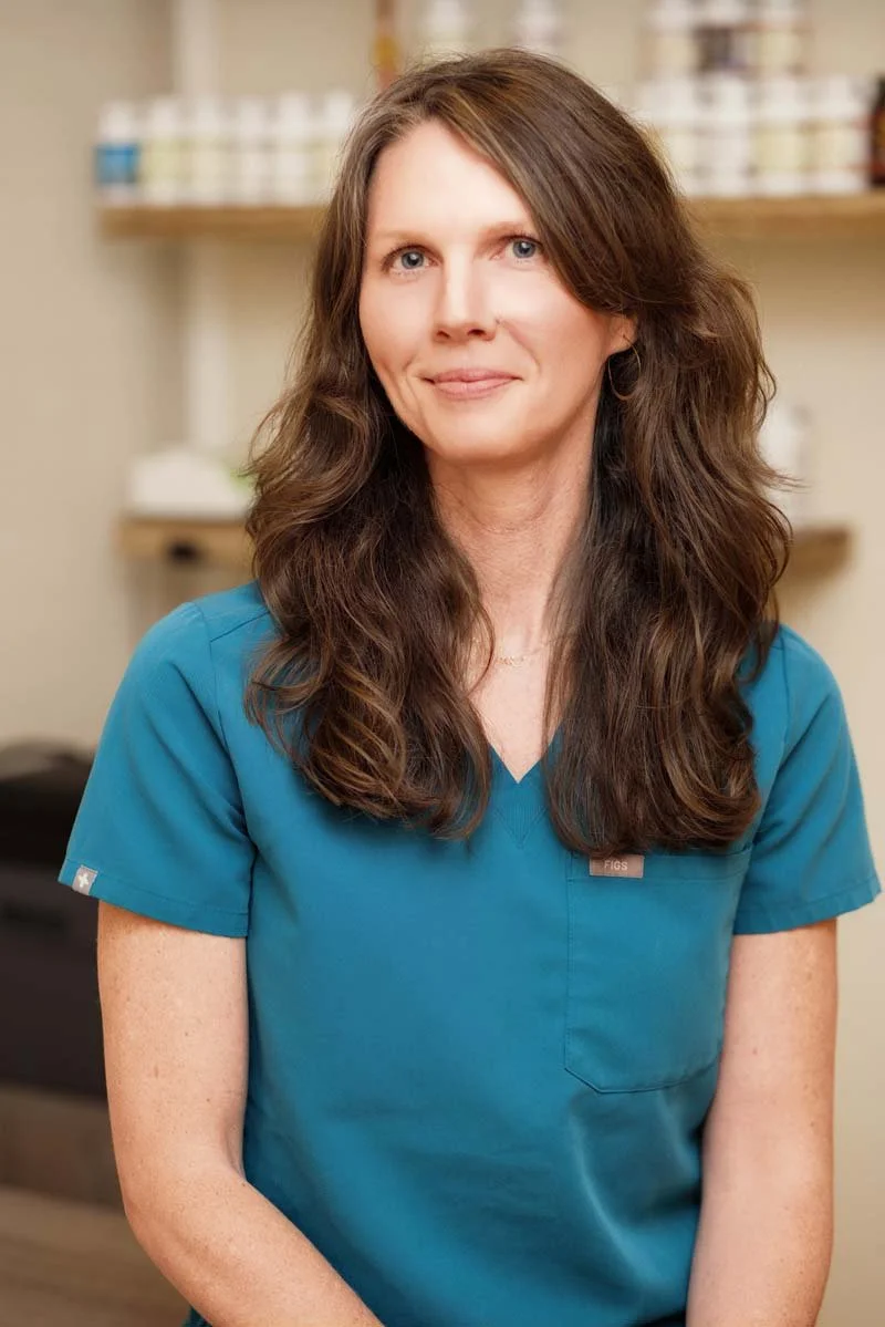 Jennifer Peterson of Current Wellness in Portland with long brown wavy hair sits, wearing blue scrubs, smiling softly, in a medical setting with shelves of bottles behind her.