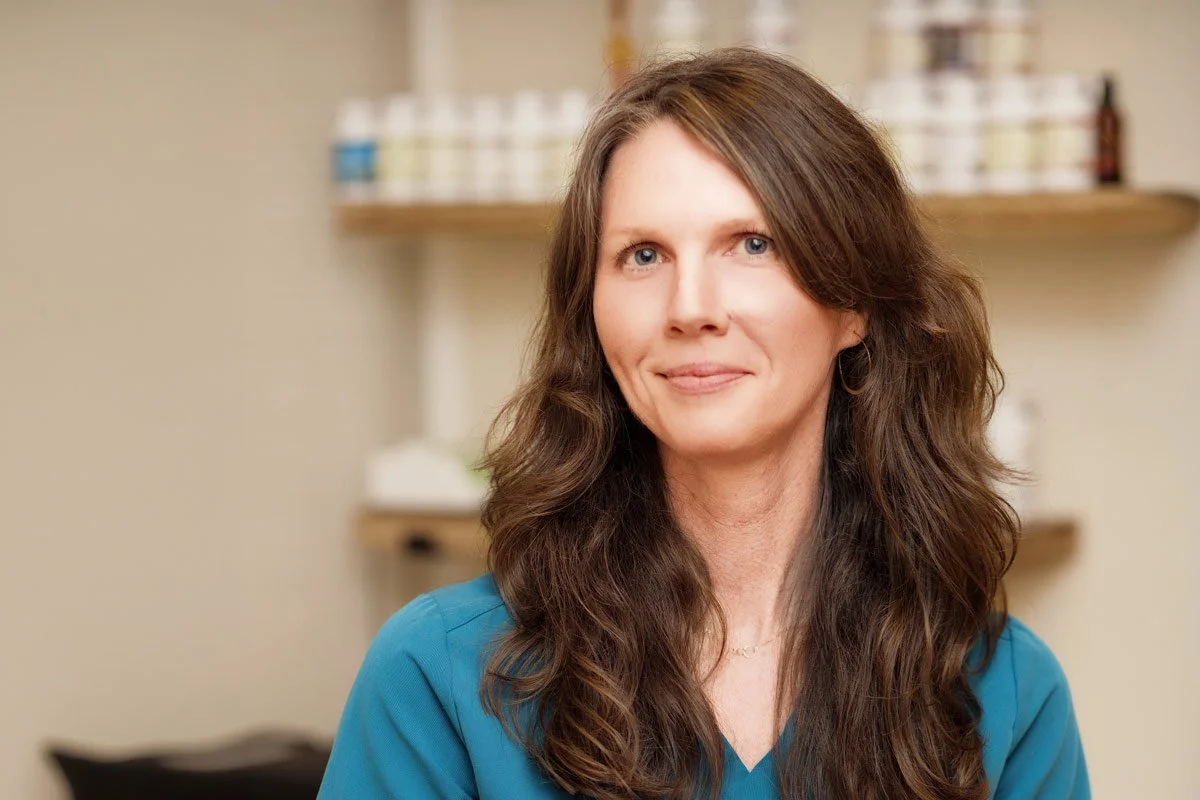 Jennifer Peterson of Current Wellness in Portland sits with long brown hair, piercing blue eyes, and light skin, wearing a blue top, smiling softly in a medical or spa setting with shelves of skincare products in the background.