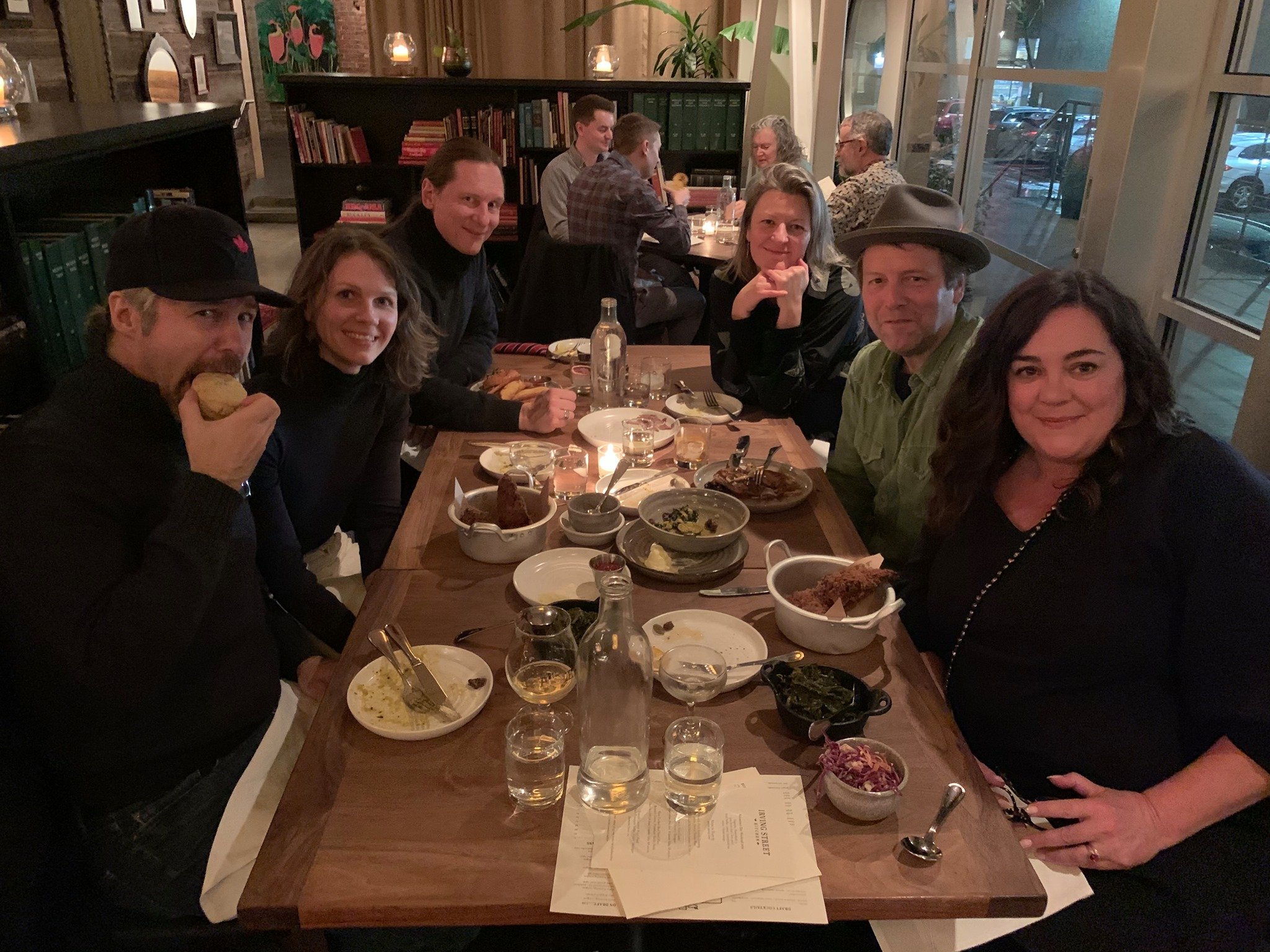 Group of eight people seated around a long wooden dinner table in a cozy restaurant, enjoying a meal together, with food and drinks visible on the table.