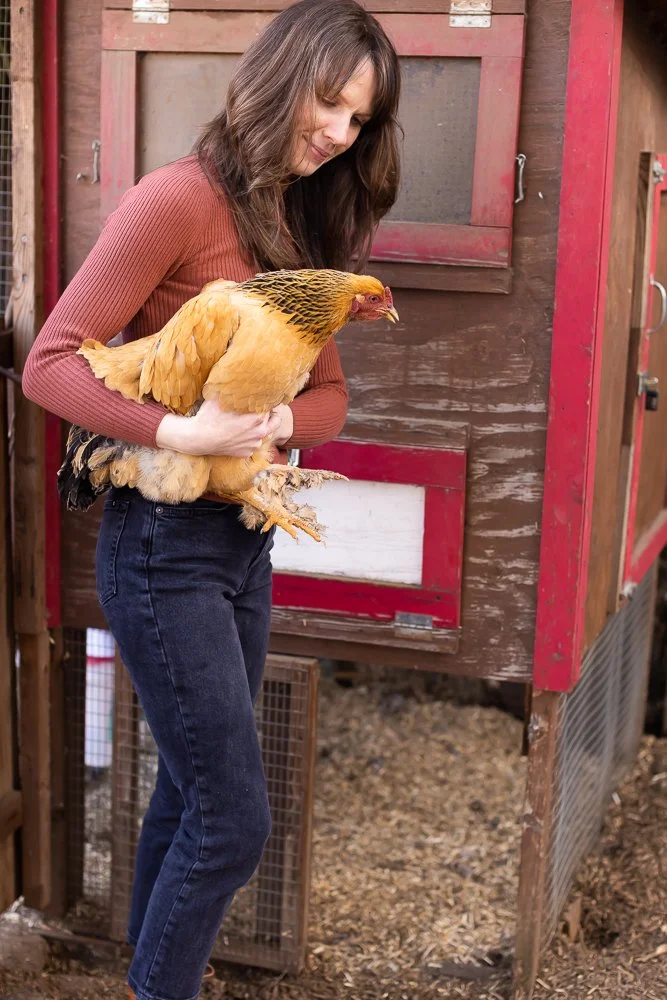 Jennifer Peterson holds a large yellow chicken in her arms, standing outside a wooden chicken coop.
