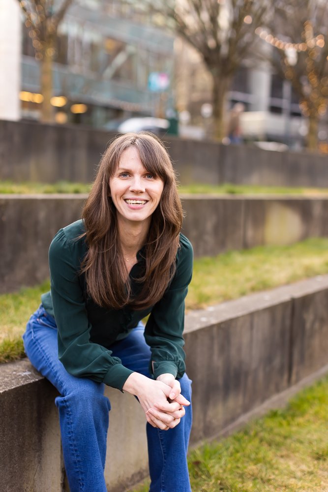 Jennifer Peterson of Current Wellness in Portland, Oregon sits on a concrete ledge outdoors, smiling at the camera, with blurred trees and buildings in the background.