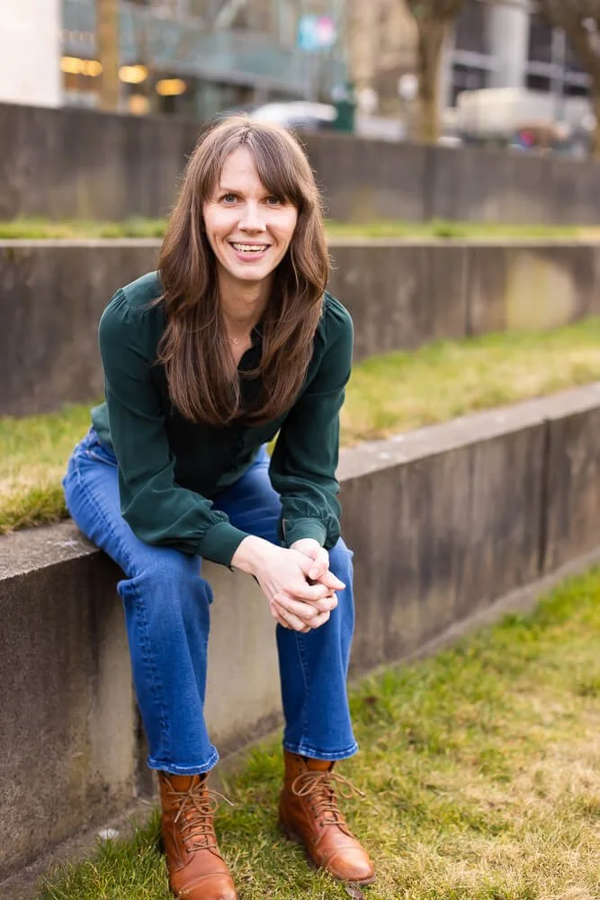 Jennifer Peterson of Current Wellness in Portland, Oregon sits on a concrete ledge outdoors, smiling at the camera, with blurred trees and buildings in the background.