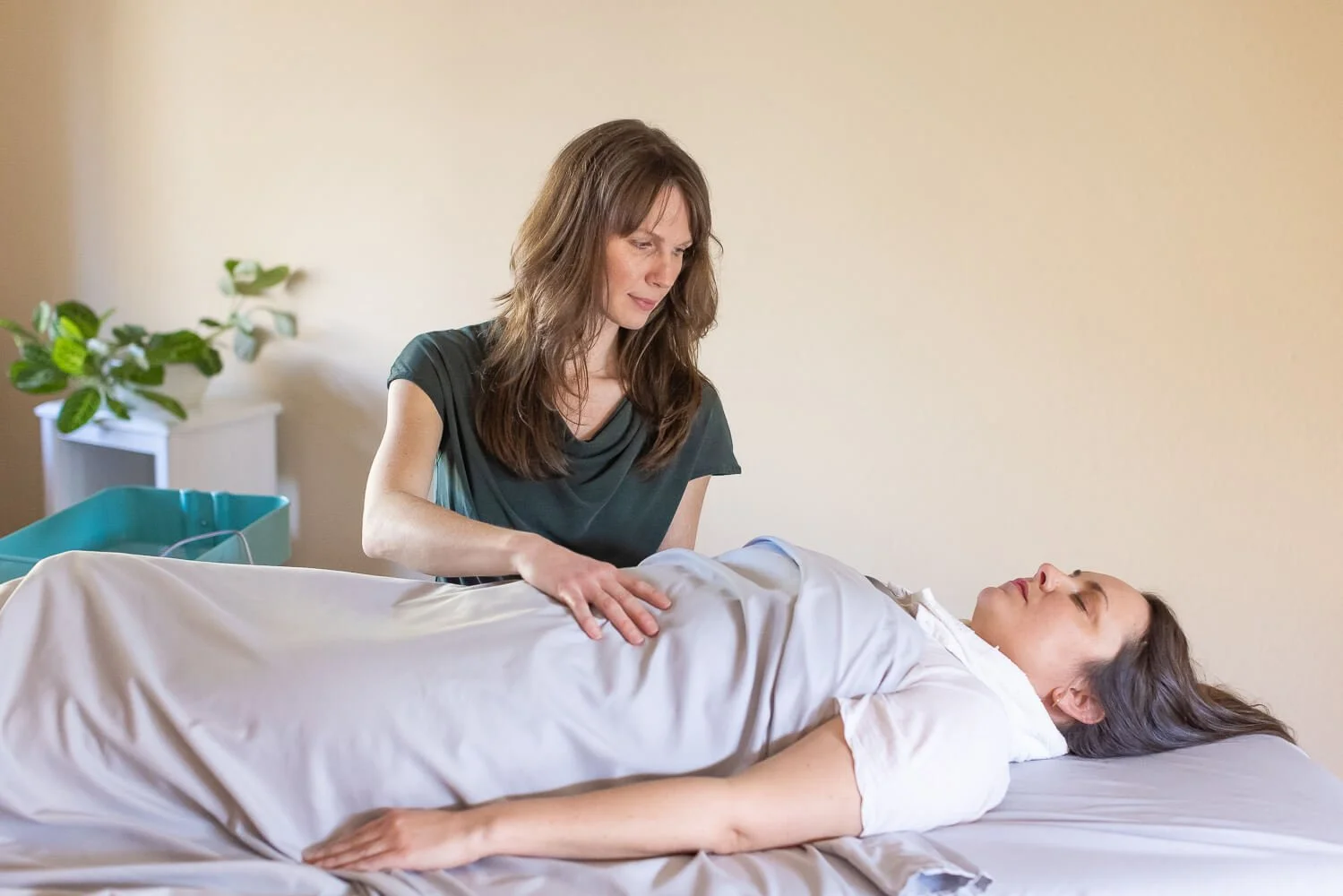 A woman is lying on a massage table with her eyes closed while Jennifer Peterson of Current Wellness in Portland, Oregon is gently holding her with one hand on her chest. She is performing manual therapy on the patient.