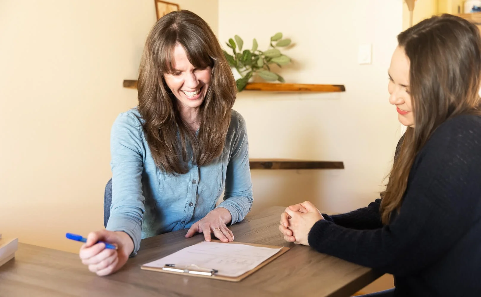 A patient with long brown hair and a blue shirt, is listening attentively to Jennifer Peterson with her arms crossed. The other woman has brown hair and a darker shirt, and is speaking or making a point. There are papers and a pen on the table.