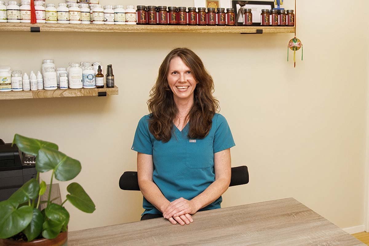 Jennifer Peterson of Current Wellness in Portland site in teal scrubs sitting at a desk in an office or clinic, smiling, with shelves of bottles and jars behind her and a plant on the desk.