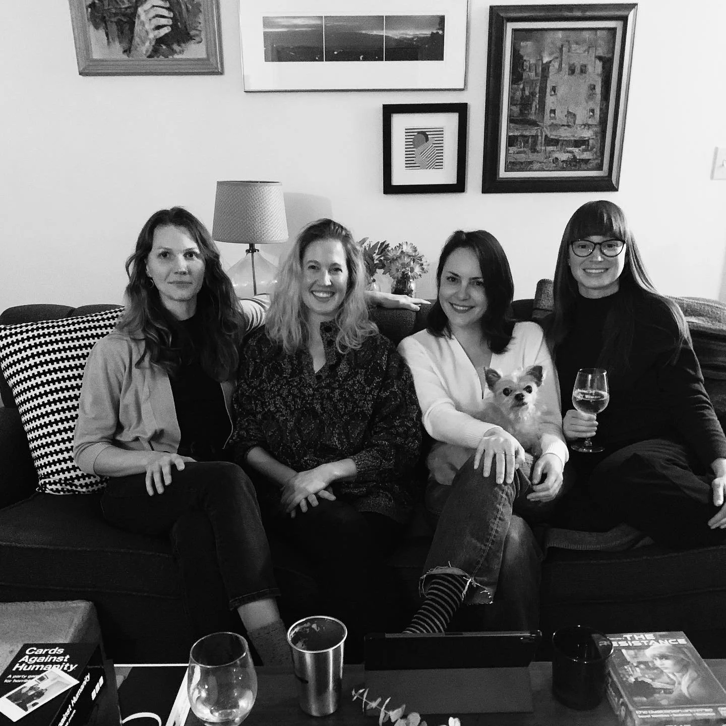 Four women sitting on a couch with a small dog, smiling. The woman second from the right is holding a glass of wine. There are framed pictures on the wall behind them, along with a table lamp.