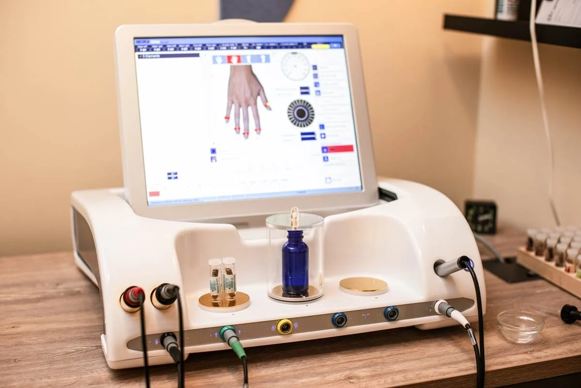 An EAV (Electro-acupuncture According to Voll) medical device at Current Wellness for testing or treatment, featuring a monitor displaying a diagram of a hand with color-coded indicators, cables connected to the device, and test tubes on the surface.